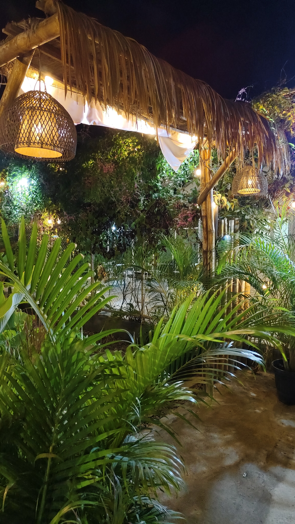The image depicts an outdoor seating area with a thatched roof structure at night. Two woven lamps hang from the structure, emitting a warm glow. The area is surrounded by lush green foliage, including palm trees and other plants. String lights are visible in the background, adding to the ambiance. The ground appears to be a mix of dirt and concrete. The scene suggests a tranquil and inviting setting, possibly a restaurant or cafe in Keudebayu, Indonesia. There are no people visible in the image.