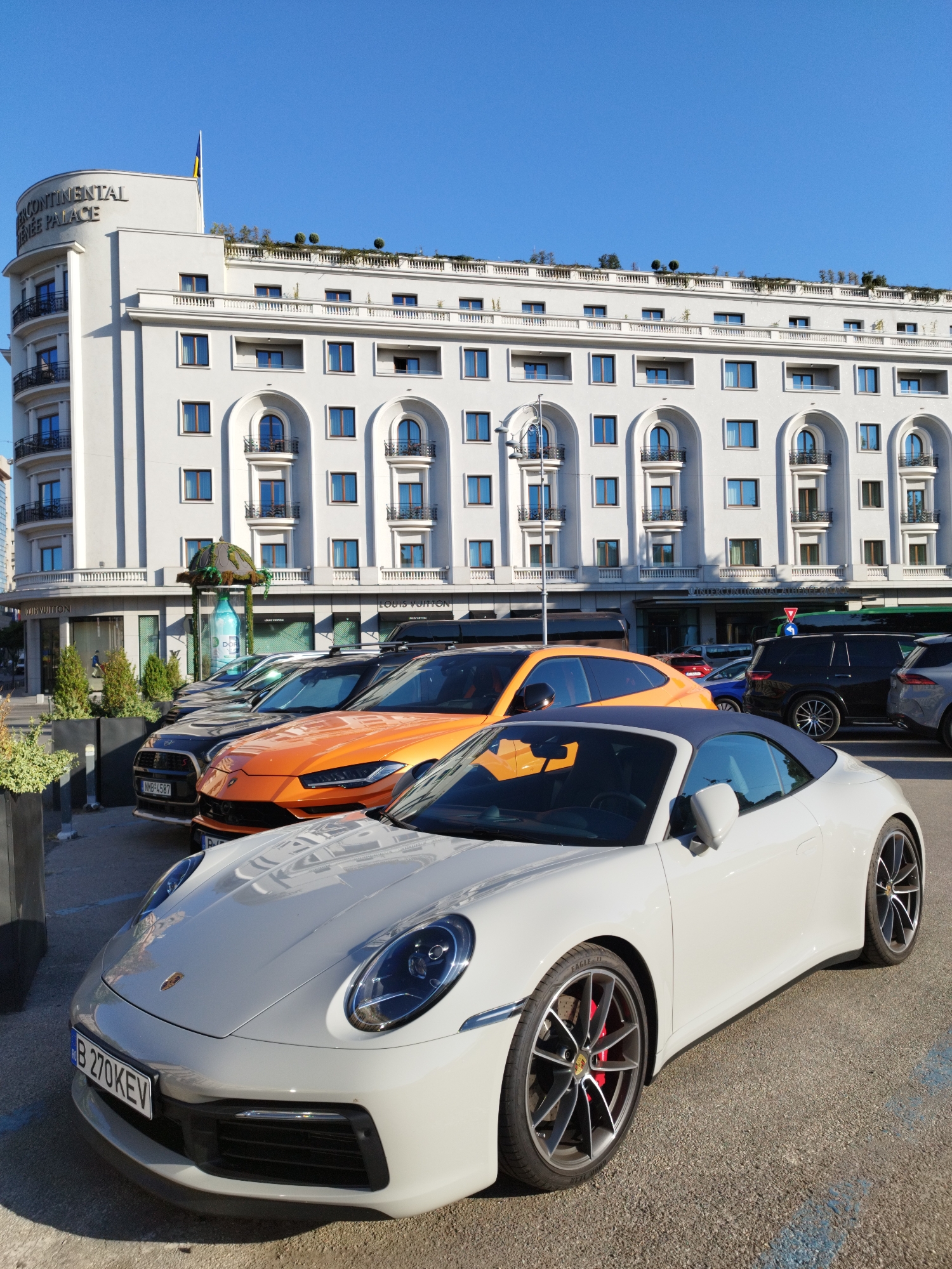 The image captures a sunny day in Bucharest, Romania, with the InterContinental Athenee Palace Hotel as the prominent background. In the foreground, a white Porsche 911 convertible is parked, its license plate displaying "B 270 KEV". To its left, an orange Lamborghini Urus SUV is parked, followed by a dark-colored Audi SUV. Behind these luxury vehicles, the facade of the historic InterContinental Athenee Palace Hotel is visible, a grand white building with arched windows and balconies. The hotel sign reads "CONTINENTAL ATHENEE PALACE". Below the hotel's arches, the Louis Vuitton boutique is identifiable, indicated by its signage. The environment is an urban setting, with paved ground and clear blue skies overhead. The overall scene suggests a high-end urban environment, likely a popular spot for luxury car owners.