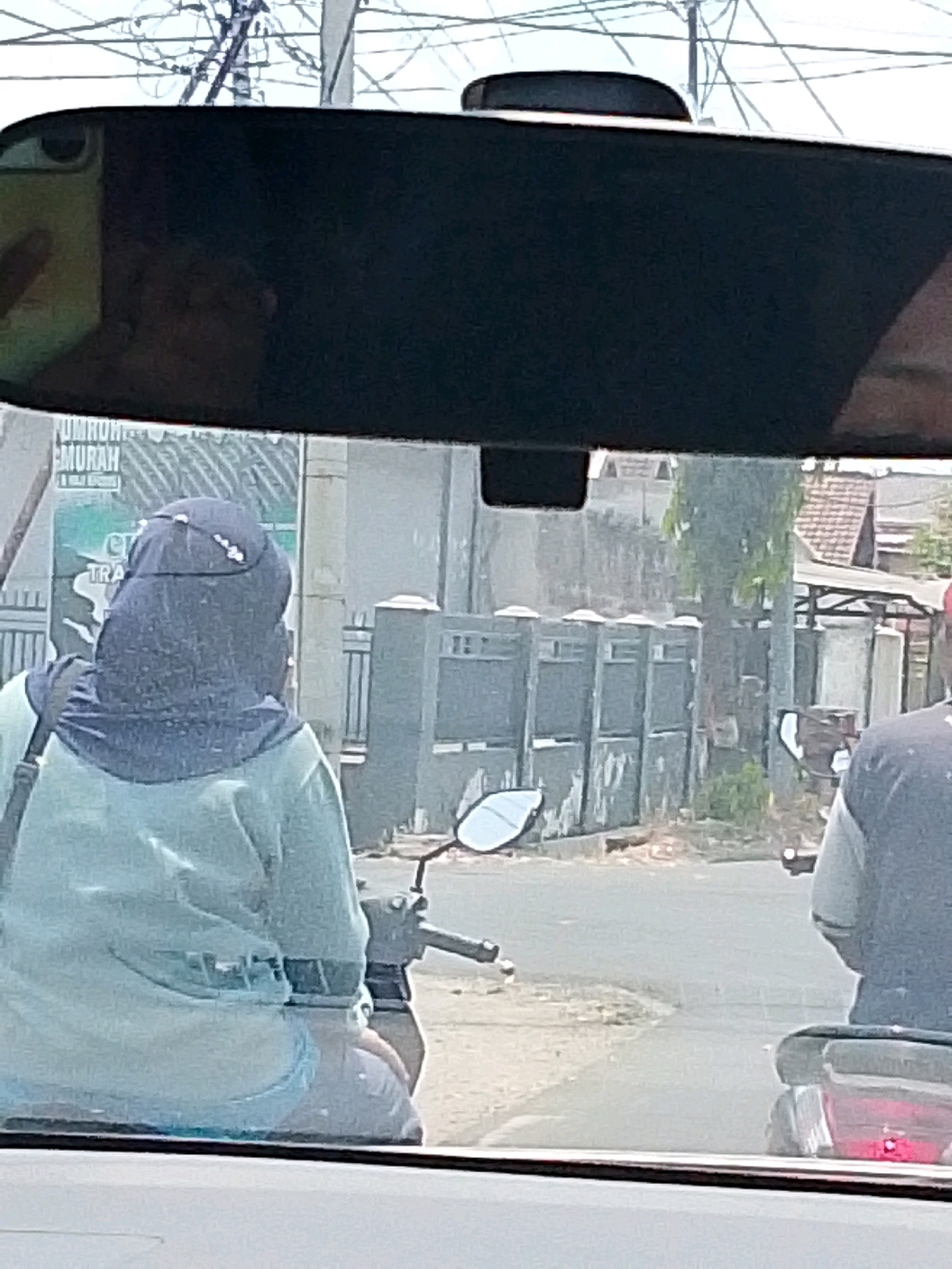 In Mojowangi, Indonesia, a person wearing a light blue long-sleeved shirt and a dark blue hijab is riding a motorcycle. The person's back is to the camera, and their head is turned slightly to the left. The motorcycle is a dark color and has a red taillight. In the background, there is a row of gray concrete pillars with metal fencing. A sign with Indonesian text is visible on the left side of the frame. Another person is riding a motorcycle in front of the main subject, also facing away from the camera. The scene appears to be a street with buildings and trees in the distance. The image is taken from inside a vehicle, likely another car, as indicated by the rearview mirror at the top of the frame.
