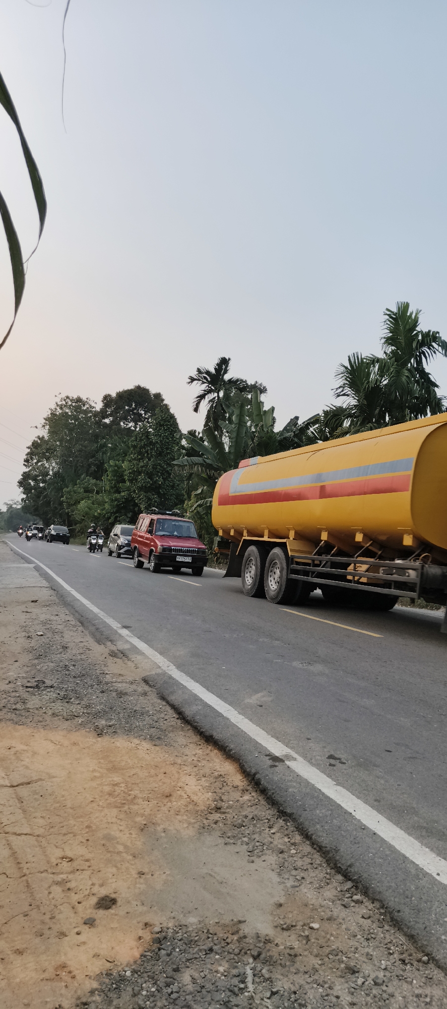 A wide-angle, eye-level outdoor shot captures a road scene in Danau Lamo, Indonesia, during daylight. The foreground on the left shows a dusty and gravelly shoulder bordering the asphalt road. The road itself is gray with a white dividing line on the left and a yellow broken line on the right. A large yellow tanker truck with red and gray stripes is prominently featured on the right side of the road, its rear wheels visible. In the distance, a line of vehicles, including a red SUV, several motorcycles, and other cars, are traveling in the same direction as the tanker truck. The background is filled with lush green trees and palm trees, suggesting a tropical environment. The sky is a pale blue with hints of orange and pink, indicative of either early morning or late afternoon. The overall impression is one of everyday traffic on a rural or semi-rural road.
