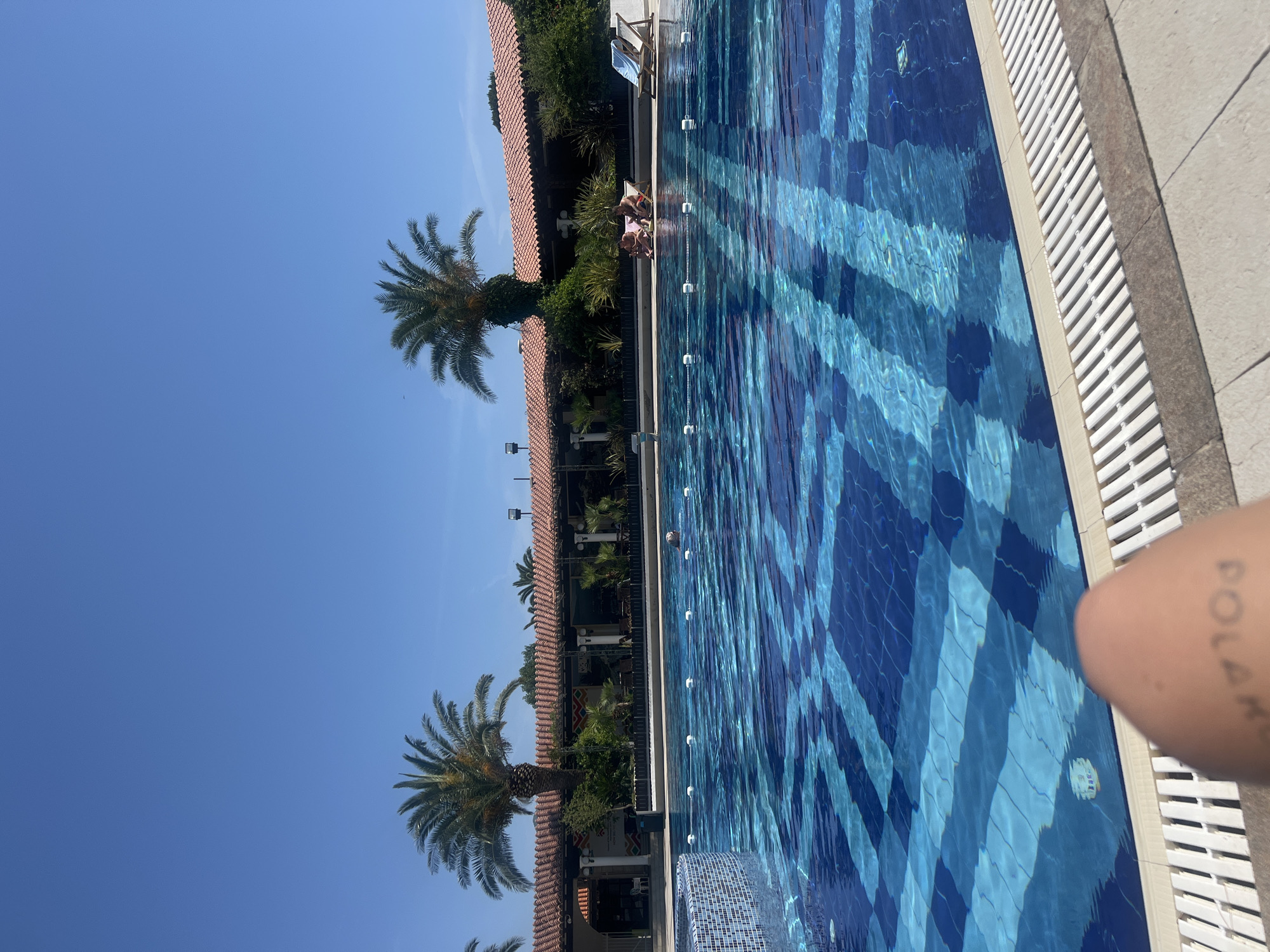 Captured poolside in Budva, Montenegro, the scene presents a partially visible tiled pool with a blue checkered pattern, extending horizontally across the frame. Alongside the pool, a building is observable, marked by a red-tiled roof and adorned with palm trees, foliage, and a series of lights or fixtures. In the backdrop, a clear, light-blue sky dominates the upper left portion. In the upper center portion of the frame, two figures are seen sitting on the side of the pool. The right side of the image reveals a fragment of a human arm with a tattoo visible.