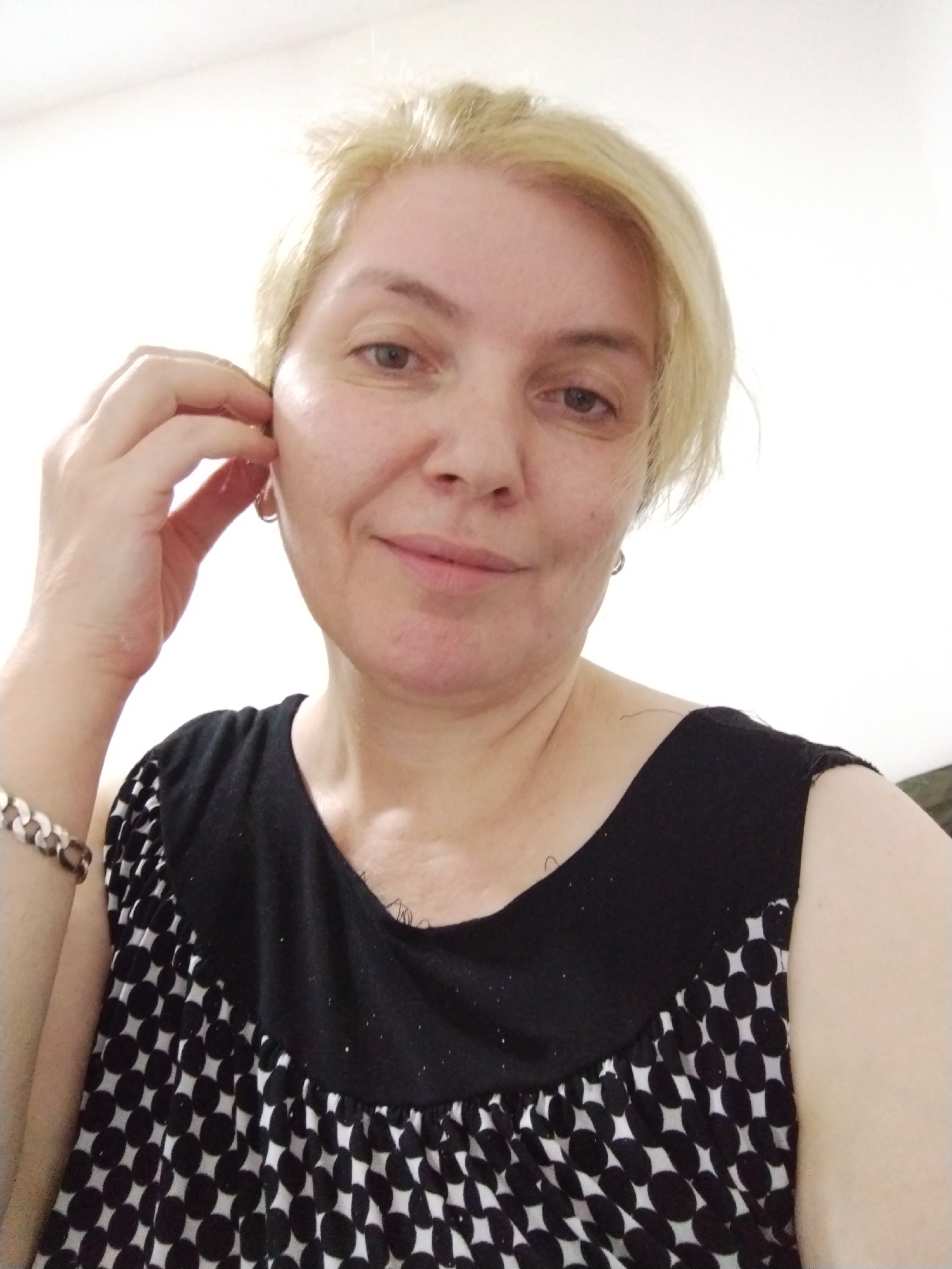 A light-skinned woman with short blonde hair is seen in a close-up shot. She is wearing a black and white patterned top. Her right hand is near her ear. A silver bracelet is visible on her left wrist. The background appears to be a plain white wall. There is no discernible information to place the image in Al Mas'oudi, United Arab Emirates.