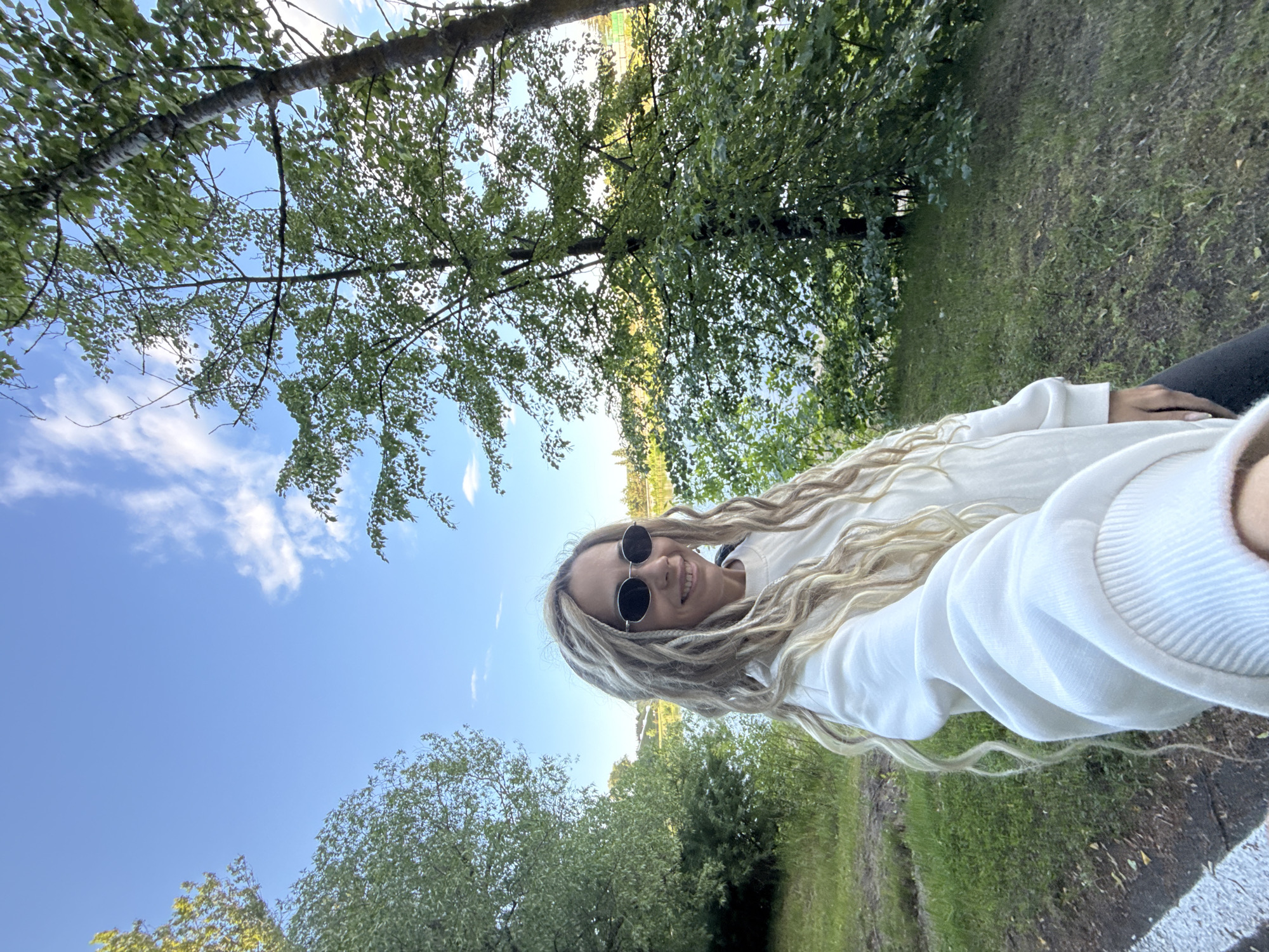 A young woman with long, light blonde hair styled in dreadlocks or thick braids is featured in a selfie, taken outdoors. She is smiling broadly and wearing dark, round sunglasses and a white long-sleeved top, with dark pants partially visible. Her arm is extended, holding the camera to capture herself and the surrounding landscape.
The setting is a vibrant natural environment, likely a park or forest edge, characterized by lush green trees and grass. A clear, bright blue sky with scattered white clouds dominates the upper left portion of the image. In the background, beyond the immediate foliage, there appears to be an open area, possibly indicating a distant field or a body of water reflecting the sunlight.
The bright natural lighting suggests it is daytime, under sunny and pleasant weather conditions. The woman's smile conveys a joyful and relaxed mood. No visible text is present in the image. The scene offers a glimpse into a serene outdoor moment, potentially in Заволжье, Russia.