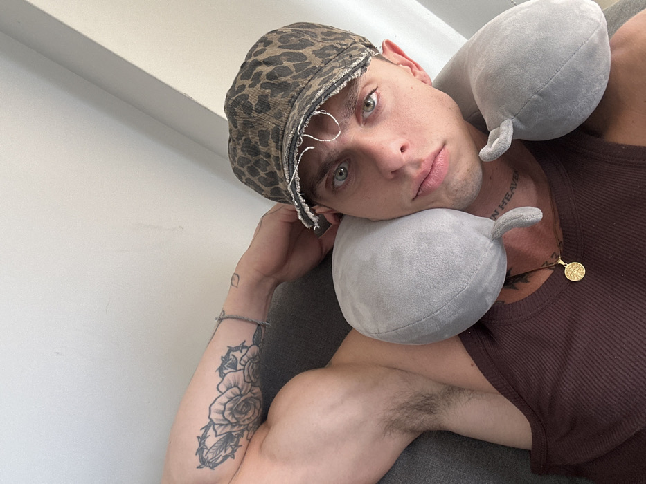 A fair-skinned person with striking blue eyes and a leopard-print cap rests their head on a grey travel pillow. They wear a brown tank top and a gold pendant necklace. Tattoos are visible on their neck ("I NEED YOU IN HEAVEN") and right arm (roses intertwined with barbed wire). A simple bracelet adorns their wrist. The background includes a light-colored wall and a glimpse of grey fabric, possibly a couch or chair. The photo is taken from a slightly high angle.