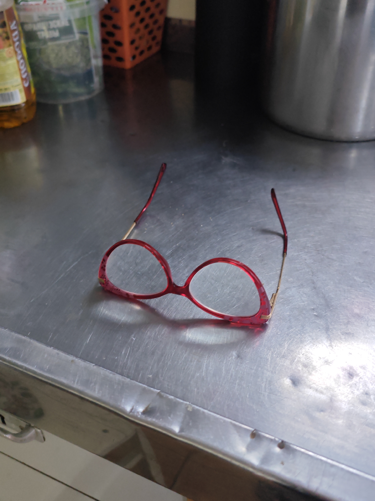 A pair of red-rimmed eyeglasses with gold temples rests on a stainless steel surface, likely a countertop. The eyeglasses are positioned slightly off-center, with the left lens and rim more prominent. The surface appears somewhat worn, with visible scratches and a few small indentations. In the background, out of focus, are what appear to be kitchen containers and possibly a metal pot. The lighting suggests an indoor environment. The location is identified as the city of Yartsevo, Russia.
