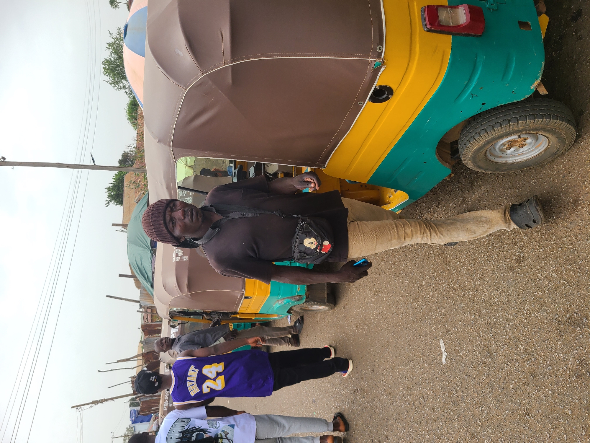 The image depicts a man standing beside a yellow and green "Keke Napep" (auto-rickshaw) on a street, presumably in Jos, Nigeria, given the context. He is wearing a dark shirt, light-colored trousers, a dark beanie, and a small black bag across his chest. He is holding a blue object, possibly a phone, in his hand. The Keke Napep has a brown roof. In the background, there are other Keke Napeps and people, including someone wearing a Lakers jersey with the number 24. The street surface is asphalt. The sky is overcast. Power lines are visible in the distance.