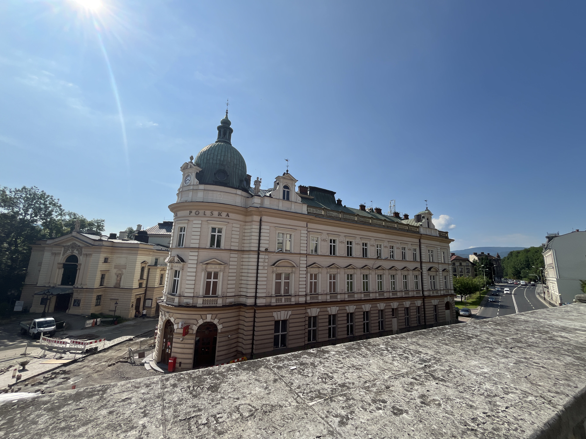 An eye-level, wide shot captures a historic building with a green dome and the inscription "POLSKA" under a clear blue sky. The building, rendered in pale yellow and beige, features numerous windows with white frames and decorative architectural elements. In the foreground, a rough, textured stone surface with graffiti occupies the bottom portion of the frame. To the left of the main building, another pale yellow structure with a grand entrance is visible, and to the far left, green trees obscure parts of the scene. Construction activity is evident in the mid-ground, with barricades, a wheelbarrow, and what appears to be paving stones laid out on a dusty ground. A white truck is parked near the construction site. To the right, a street with cars and more trees stretches into the distance, with mountains visible on the horizon under the bright sun, which creates a lens flare in the upper left quadrant.