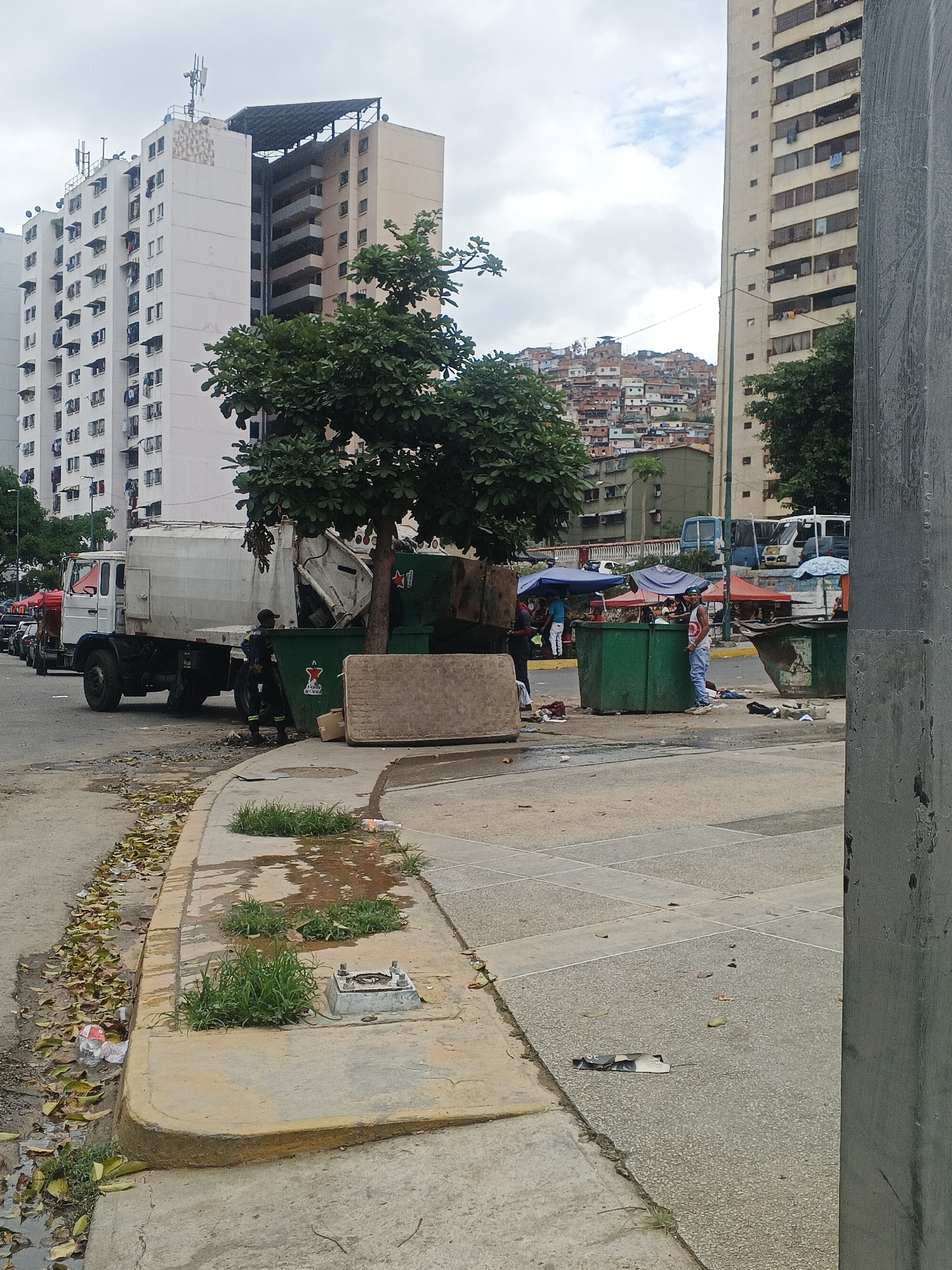 The scene depicts a street in Caracas, Venezuela, with a garbage truck parked beside overflowing green dumpsters. A sanitation worker stands near the truck. A mattress and other debris are scattered around the dumpsters. People are seen standing by the dumpsters, possibly vendors under tents. Buildings are in the background, with one prominent, light-colored high-rise apartment building. The curb is stained with water and debris, and a metal access point is visible. The background shows a hillside densely populated with buildings. The sky is overcast.