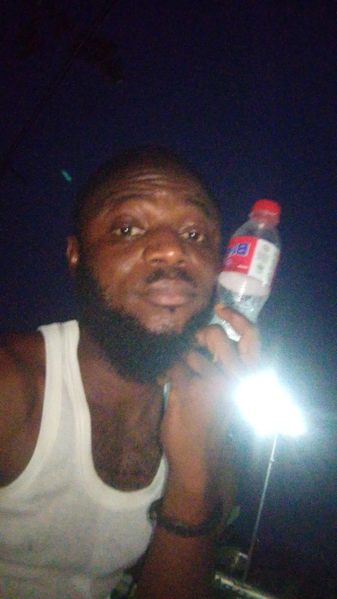 A man is holding a plastic bottle of water in front of a dark background. The man has a beard and is wearing a white tank top. The location in Lokoja, Nigeria, is unknown.