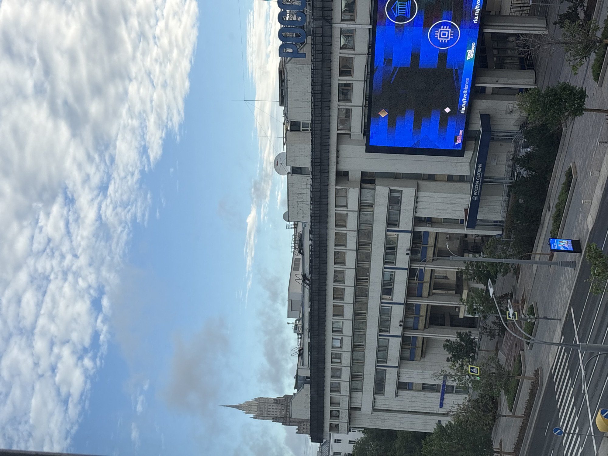 The image captures an urban scene in Moscow, Russia, dominated by a large, brutalist-style building under a cloudy sky. A prominent spire of a Stalinist skyscraper, likely the Ministry of Foreign Affairs building (Smolenskaya Square, 32/34), is visible in the upper left background, partially obscured by clouds. The main building in the foreground features a long, rectangular facade with multiple floors of windows. A large, bright blue LED screen is mounted on the right side of the building, displaying digital content with logos and text, including "РОССИЯ СЕГОДНЯ" (Russia Today) and "ria.ru/tv/oibiznes". Below the screen, the entrance to the "РОССИЯ СЕГОДНЯ" building is visible, with stairs leading up to a dark entryway. The street level in front of the building shows a modern urban design with trees, pedestrian walkways, and a crosswalk. Traffic lights and street signs are also present. The overall atmosphere is that of a typical cityscape with a focus on architecture and media.