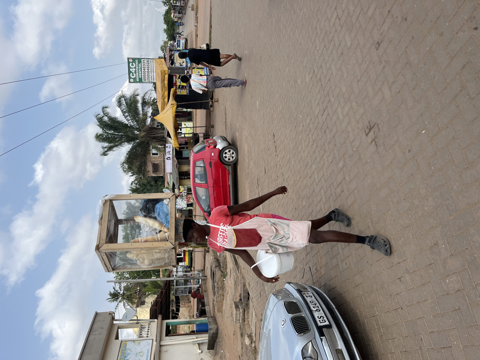 This image captures a street scene in Adenta Municipality, Ghana. In the foreground, a dark-skinned individual, wearing a black cap, red t-shirt, pink shorts, grey Crocs, and a white apron with pockets, walks from left to right. This person balances a large, rectangular wooden display case containing multiple baguettes and other items on their head, while holding a white plastic bucket in their left hand.
To the immediate left, the front portion of a silver BMW sedan with license plate "GS 6149 Z" is parked. The ground in the foreground and mid-ground is paved with light brown, herringbone-patterned bricks.
In the mid-ground, a red compact car is parked. Two additional individuals are seen walking away from the viewer on the paved surface further down the street. The background consists of various structures and vegetation. To the left, a light-colored building features a satellite dish and a blue barrel at its base. Towards the center-right, a yellow tented stall is visible next to a green sign for "C4C HOMEOPATHIC," displaying phone numbers (0500040331, 0500040328) and a website (WWW.ADURICALS.COM). Numerous trees, including a prominent palm tree, and other buildings line the street. The sky overhead is partly cloudy, and power lines are visible across the upper portion of the frame.