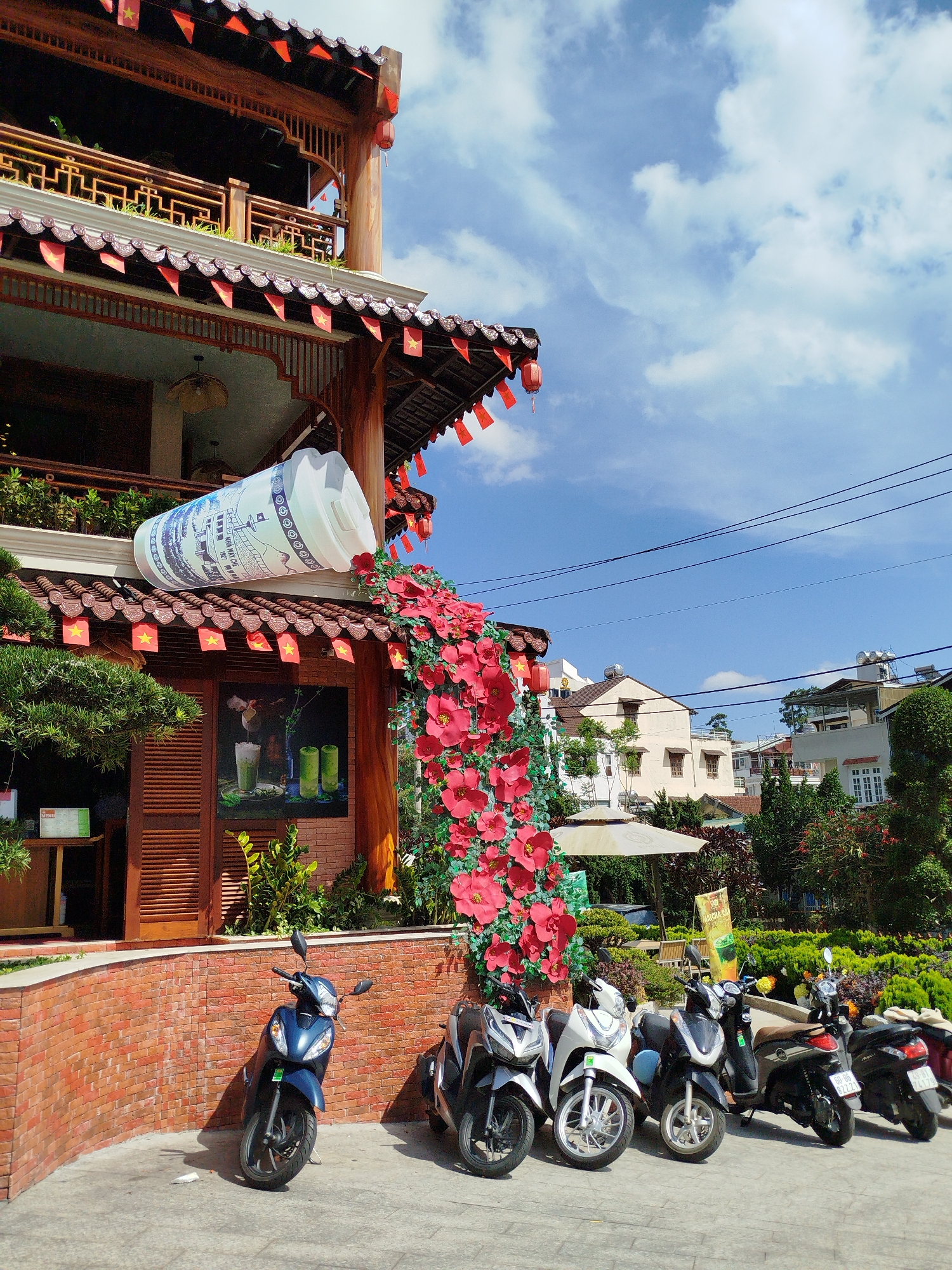 This exterior shot, taken in Cam Ly - Đà Lạt, Vietnam, depicts a multi-storied building decorated with Vietnamese flags. An oversized, cylindrical advertisement resembling a milk tea container is mounted on the roof, appearing to spill red flowers down the side of the building. A row of parked motorbikes lines the front of the building on a concrete surface. The sky is partly cloudy with power lines visible in the background. Other buildings and greenery are visible across the street.