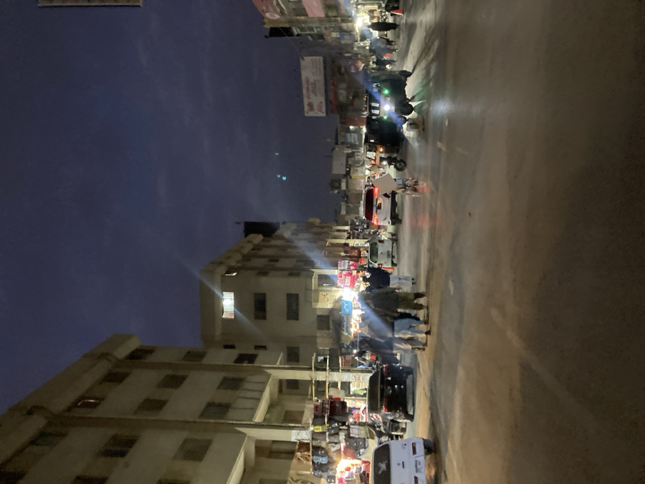 The image depicts a nighttime street scene in Quetta City Tehsil, Pakistan, under a dark, cloudy sky. The environment is urban and commercial, illuminated primarily by artificial light sources.
On the left, a multi-story, light-colored building with numerous windows occupies the foreground and midground. Its ground floor hosts multiple open-front shops displaying merchandise, possibly bags or textiles, under bright lights. Further down, similar commercial buildings line the street on both sides, featuring various signs and storefront illumination. A single window on an upper floor of a building in the midground emits a bright white light.
The paved road appears somewhat reflective, suggesting moisture or strong light reflection. Vehicle and pedestrian traffic is present. A white hatchback is parked or moving slowly on the far left. A dark sedan is positioned behind it. In the midground, a group of approximately three to five men, dressed in traditional attire, are walking on the road towards the viewer. A white SUV with illuminated taillights is further down the road on the right. Two green auto-rickshaws with headlights on are visible further into the intersection or street. A person on a bicycle, possibly carrying a large white item, is positioned between the pedestrians and the auto-rickshaws.
Pedestrians are also visible on the sidewalks and further down the street. Streetlights, shop lights, and vehicle head/taillights contribute to the scene's illumination. Distant green lights are observable against the dark sky, possibly traffic signals or building lights.