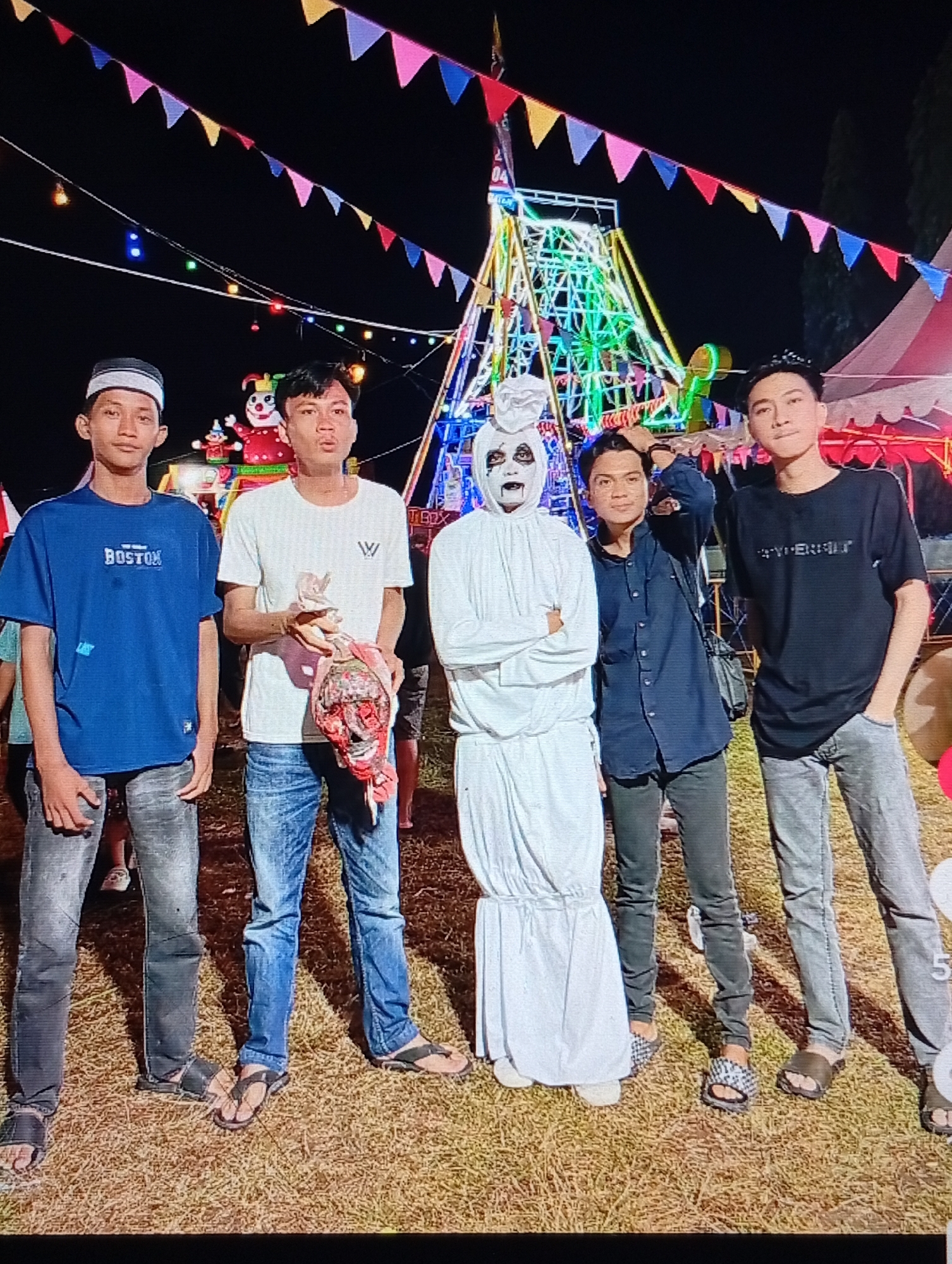 An outdoor nighttime photograph captured in Lhokseumawe, Indonesia, features five individuals posing in front of a brightly lit amusement park or carnival setting. The ground consists of dry, sparse grass or dirt. Overhead, multiple strands of colorful triangular bunting flags and string lights illuminate the dark sky.
From left to right:
1. A young male wears a white and black kufi (cap), a blue short-sleeved t-shirt with "BOSTON" lettering on the chest, faded dark jeans, and dark open-toed sandals. He stands with his hands at his sides, looking directly at the camera with a neutral expression.
2. A young male in a white short-sleeved t-shirt with a dark "W" logo, blue jeans, and dark open-toed sandals holds a red and white tattered object, resembling a dismembered doll or prop, in his left hand at waist level. He looks towards the camera with a slightly open mouth.
3. A central figure is dressed in a full-body white hooded costume, characteristic of a pocong, with its visible face painted white, dark hollowed-out eyes, and a dark wide-open mouth. Its arms are crossed over the chest, and it wears white footwear.
4. A young male wears a dark blue long-sleeved button-down shirt with sleeves rolled up, dark trousers, and black and white checkered open-toed slides. His right hand is positioned at the back of his head, and a dark strap, possibly from a bag, crosses his chest. He looks towards the camera with a slight smile.
5. A young male in a black short-sleeved t-shirt with light text on the chest, faded grey jeans, and dark open-toed sandals stands with his right hand in his pocket. He looks towards the camera with a neutral expression and a faint smile.
In the background, a large Ferris wheel-like ride with green and yellow lights is prominent in the center-left. To its right, another illuminated ride structure and two large, colorful clown figures are visible. A red and white striped carnival tent is situated on the far right, and dark tree foliage is present against the night sky.