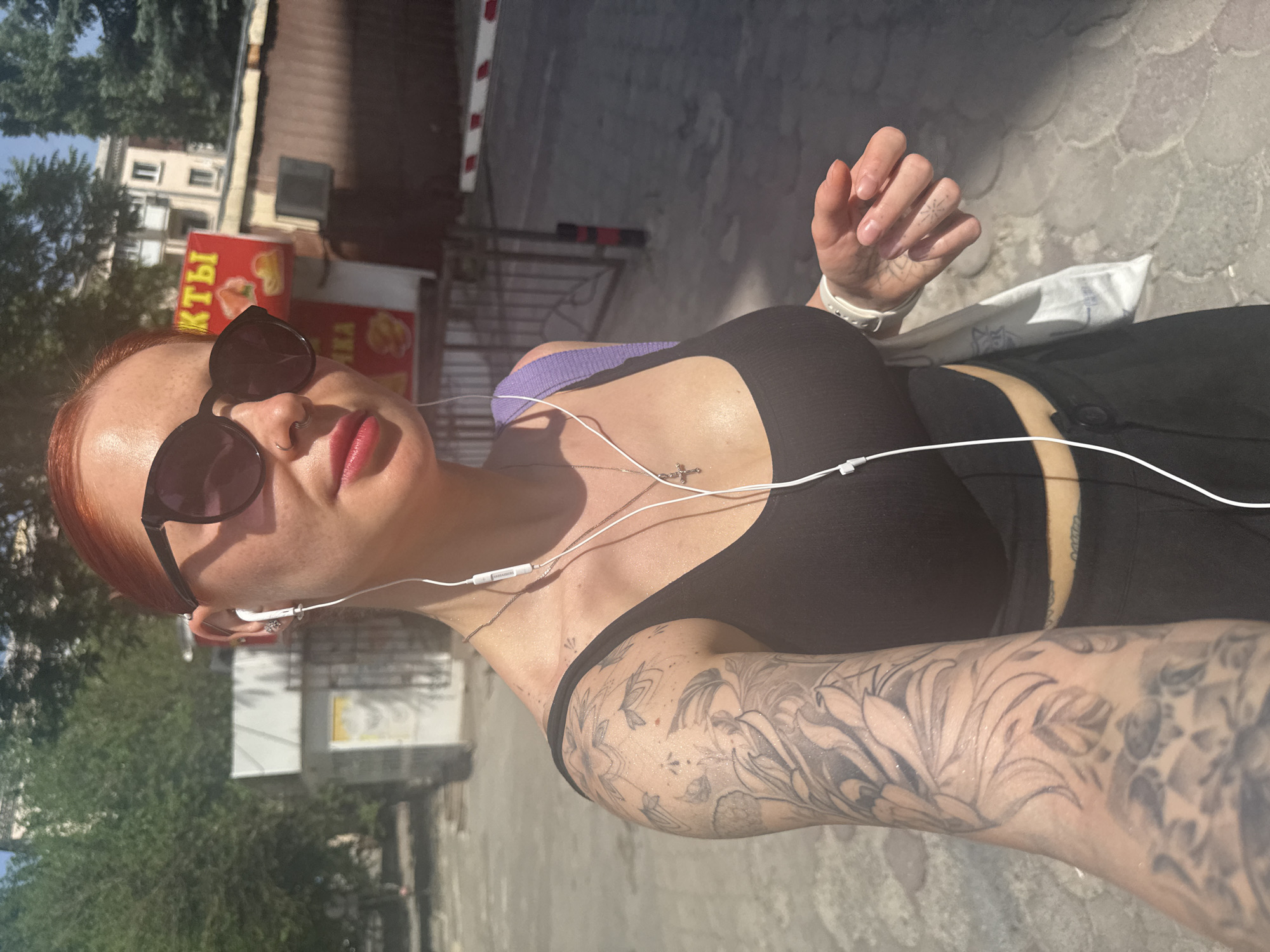 A selfie taken in Rostov-on-Don, Russia shows a fair-skinned person with red hair, sunglasses, and red lipstick. They are wearing a black tank top, headphones, and a smartwatch. Tattoos are visible on the arm and chest. The background includes a building with a sign in Russian, trees, and a paved street.