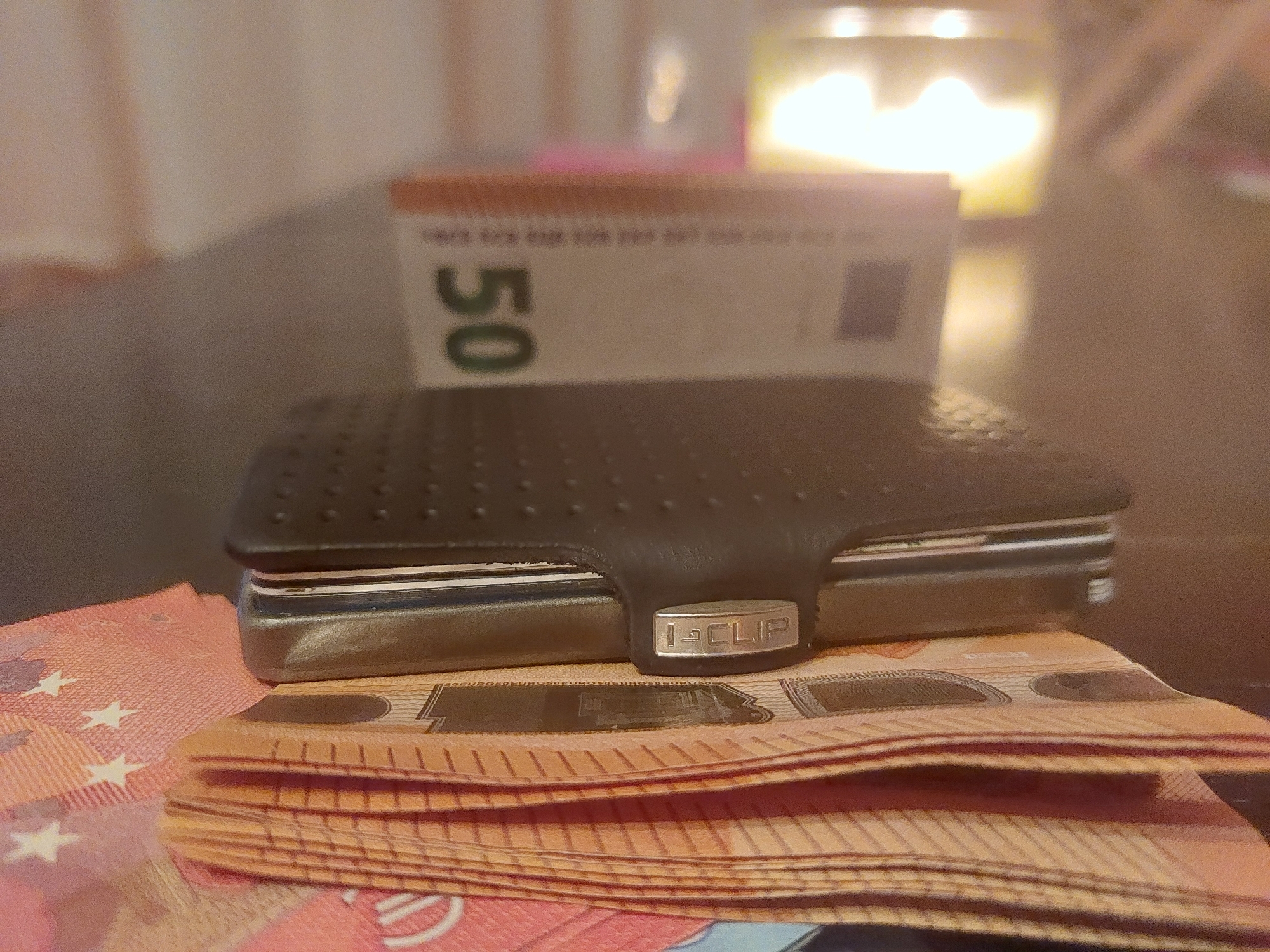 A black, metal, "I-CLIP" wallet rests on a stack of orange 50 Euro banknotes. Another 50 Euro banknote stands upright blurred behind the wallet. A blurred light source is in the upper right. The scene is indoors. There is no information to suggest it is located in Tsarevo, Bulgaria.