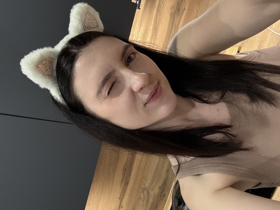 A young woman winks at the camera, wearing a fluffy white headband with cat ears. She has dark hair, fair skin, and is wearing a tan tank top. The background includes a dark gray wall and light wood paneling. The image appears to be a selfie taken indoors with natural lighting.