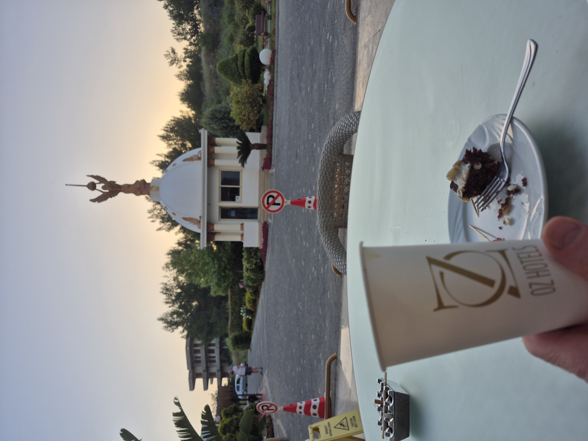Image taken from a cafe in Alanya, Turkey. Foreground shows a white table with a wicker chair, a plate with half-eaten cake, a fork, an ashtray, and a paper cup with the logo "OZ HOTELS". A hand is holding the cup. In the background, there is a building with a white dome and a statue of an eagle on top, surrounded by trees and bushes. A person can be seen walking in the distance. The sky is bright with sunset.