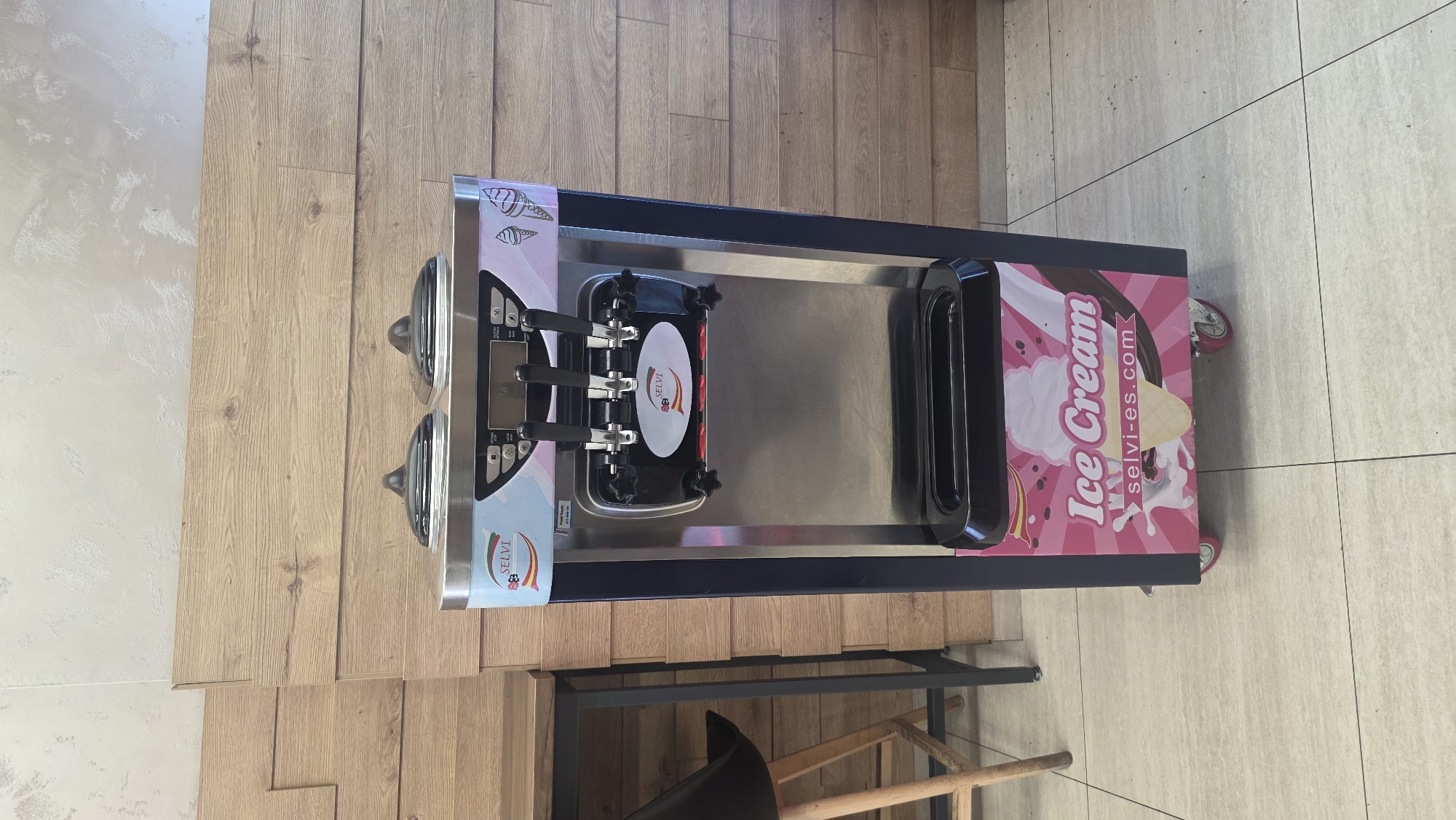 The image shows a Selvi brand ice cream machine. It is a stainless steel machine with black handles, a dispensing area, and a pink and white graphic design on the side that says "Ice Cream" and "selvi-es.com." The machine has four caster wheels for mobility. It is located on a wooden-planked surface next to a tiled surface. Beneath the ice cream machine is a black metal table with a wooden chair in the lower portion of the image. This is likely an indoor commercial setting, but the exact location in Burgas, Bulgaria cannot be determined from the image alone. There are no people visible in the frame.