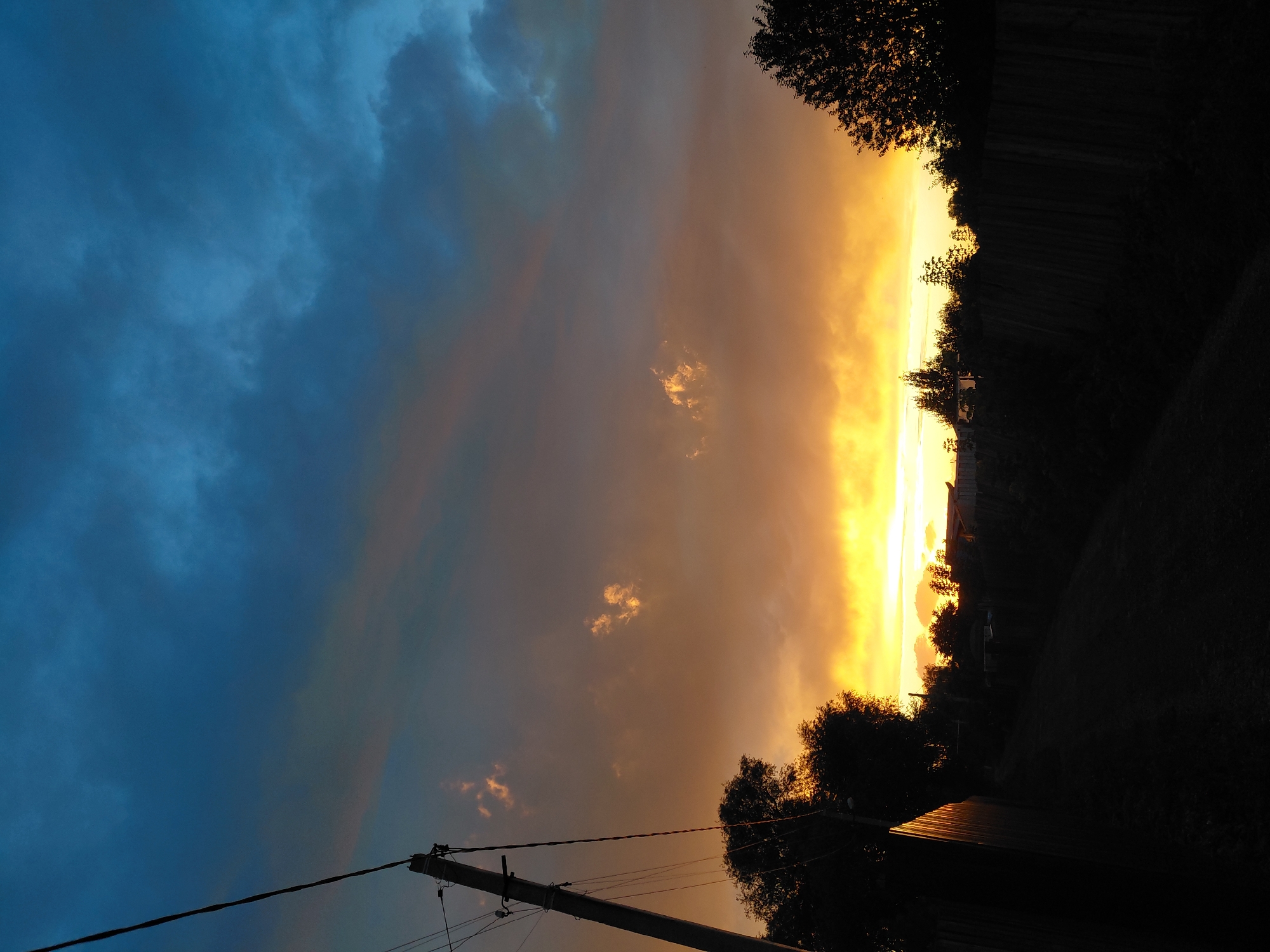 This image depicts a sunset in a rural or suburban area, likely in Novosibirsk, Russia. The sky is a dramatic gradient of colors, transitioning from a deep blue at the top to vibrant oranges and yellows near the horizon. Wisps of clouds are scattered throughout, catching the light of the setting sun.
In the foreground, a wooden fence runs horizontally across the lower portion of the frame, casting a dark shadow. To the left, a utility pole with electrical wires is prominent, silhouetted against the colorful sky. Trees and the rooftops of low-rise buildings are visible behind the fence, also silhouetted and appearing as dark shapes. The ground in the foreground is dark and appears to be a dirt path or unpaved road.
The overall impression is one of a tranquil evening scene in a less urbanized setting, characterized by the striking natural beauty of the sunset.