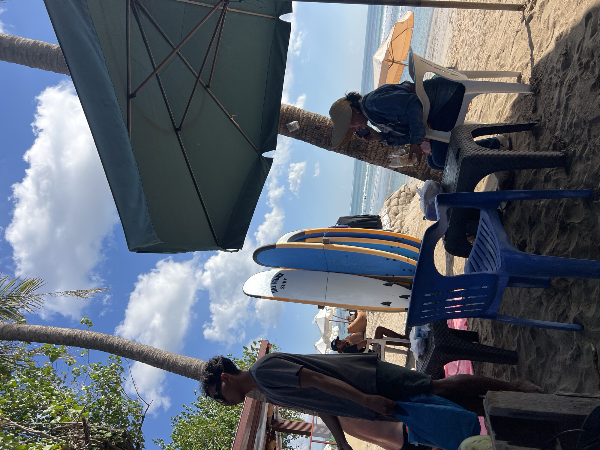 At a beachside establishment in Kuta, Indonesia, a man wearing sunglasses and a dark t-shirt stands near a table, holding a blue bag. Two surfboards, one white with "BALIGUNS SURF" written on it and a blue and orange one, are propped up nearby. A woman sits in a white plastic chair under a large green umbrella, wearing a denim jacket and a hat, and is speaking on her phone while holding a bag of food. She is seated at a small black table. In the background, the ocean waves gently roll onto the sandy shore, and several beach umbrellas are visible. The scene is illuminated by bright sunlight, casting shadows on the sand.