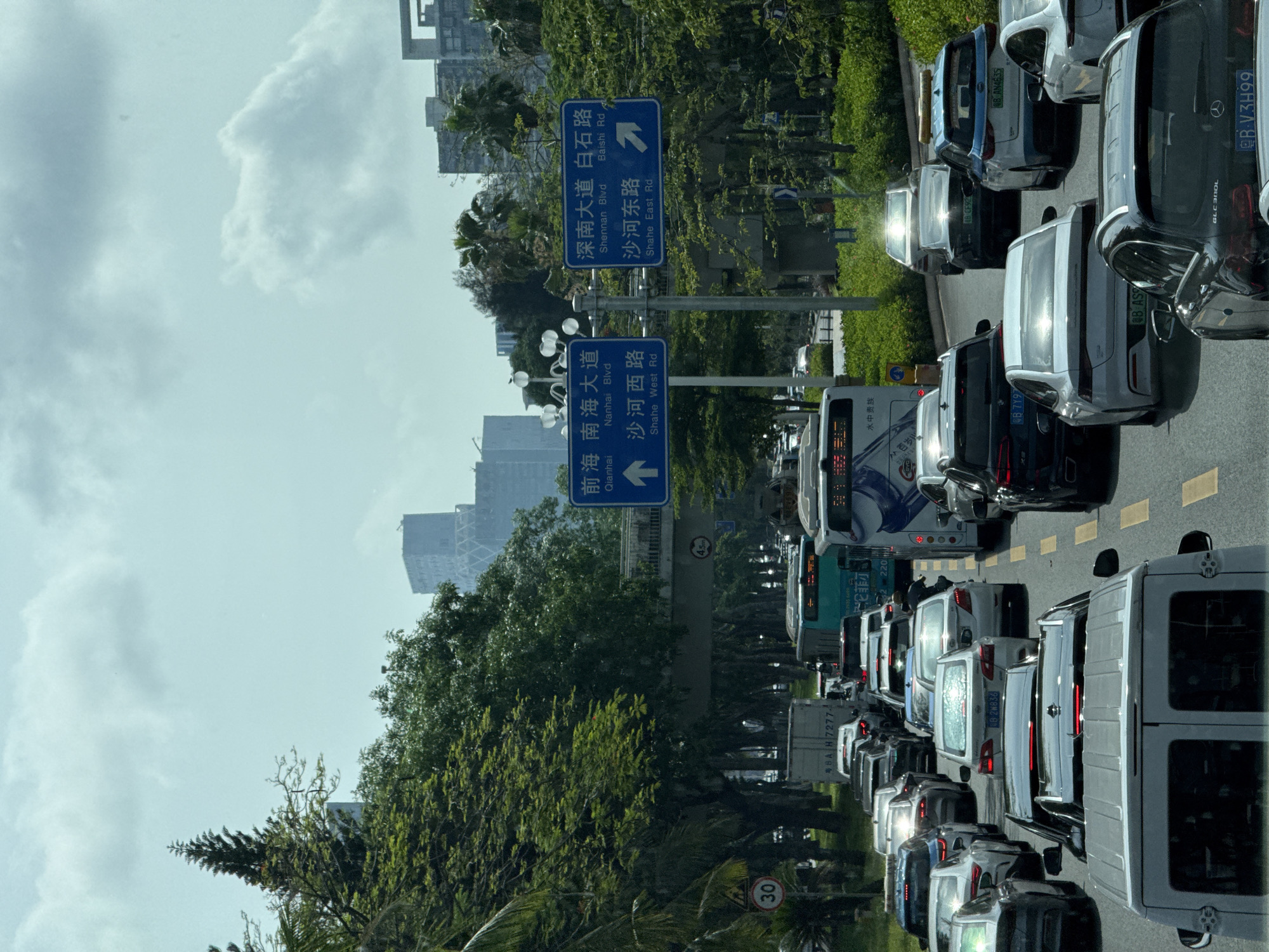 The image depicts heavy traffic in Nanshan District, Shenzhen, China. The traffic jam is on a multi-lane road with yellow lane markings. Road signs indicate directions to Qianhai, Nanhai Boulevard, Shahe West Road, Shennan Boulevard, Baishi Road, and Shahe East Road. The surrounding environment includes trees and buildings, suggesting an urban setting. Vehicles of various types, including cars, buses, and vans, are visible in the traffic. The sky is overcast with visible clouds.