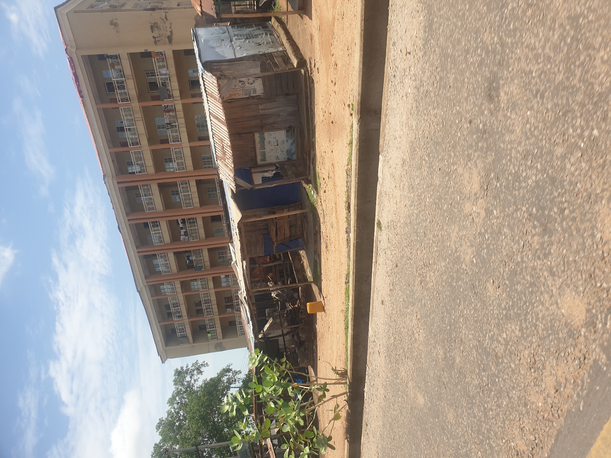 The image shows a street scene in an urban area, likely in Unknown, Unknown. A multi-story apartment building stands prominently on the left. Adjacent to it, a makeshift wooden structure serves as a roadside stall. The stall is constructed from wooden planks and corrugated metal, and has advertisements posted on its exterior. A small yellow container sits near the base of the stall. A narrow, unpaved area separates the stall from the road. The road itself is asphalt and occupies a large portion of the right side of the frame. A small green bush is visible in the foreground. The sky is partially visible with some clouds.