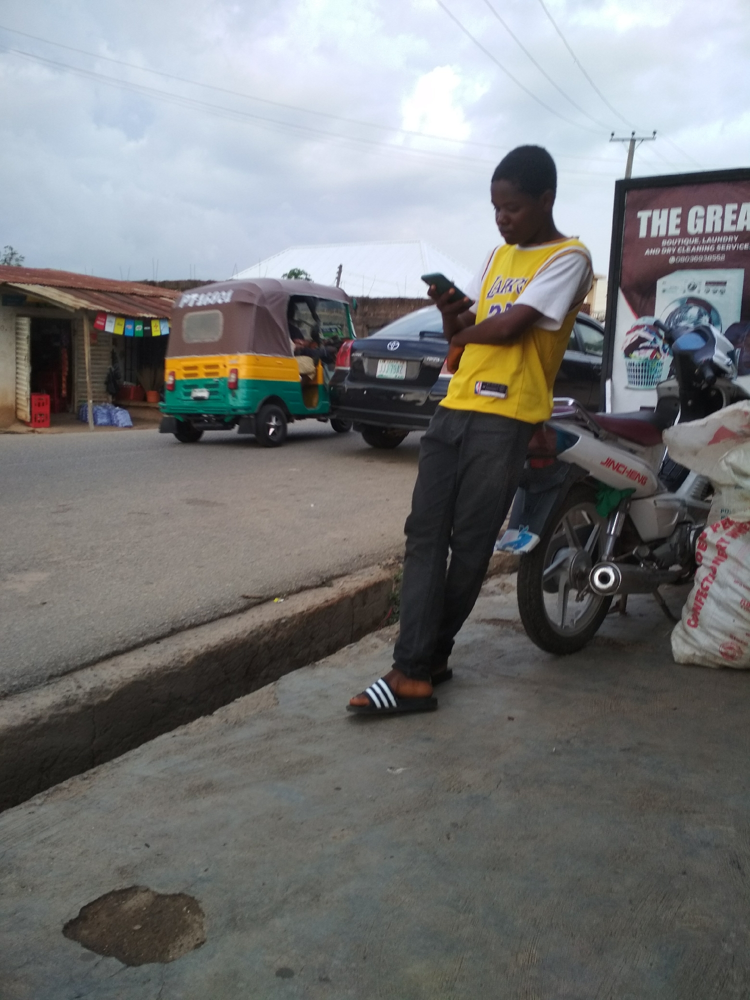 A young man is standing on a sidewalk, leaning against a motorcycle. He is wearing a yellow jersey, dark jeans, and Adidas slides and is looking at his phone. Behind him is a black Toyota car and a green and yellow tricycle, parked on the street. A small shop with colorful decorations is visible across the street. A large sign that reads "THE GREAT" is also visible near the motorcycle. The sky is cloudy. The location is a city identified as Unknown, Unknown.