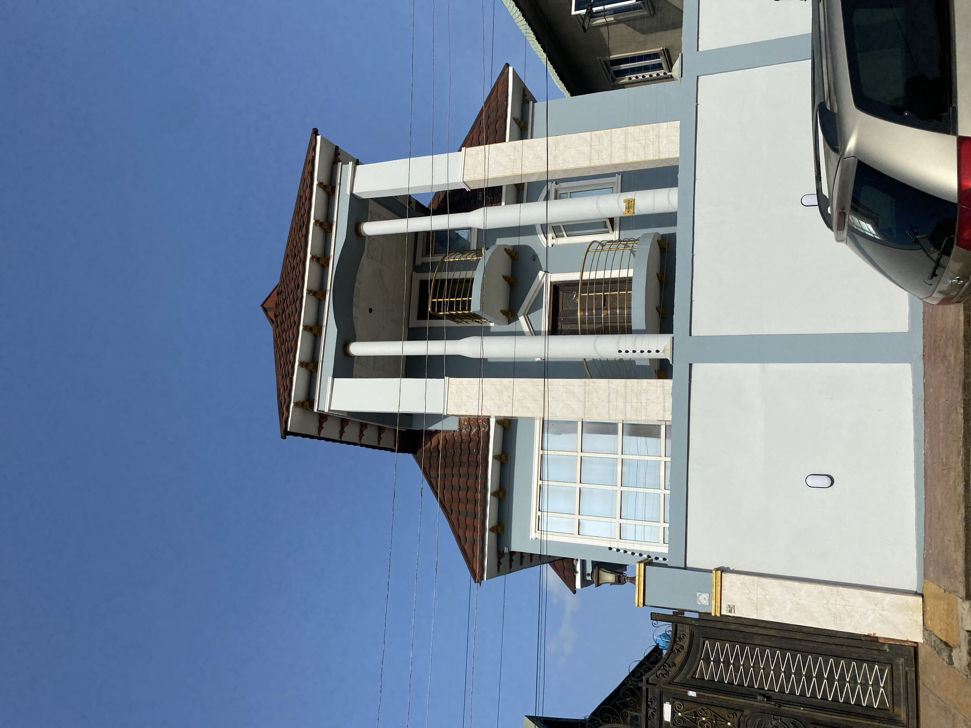 The image depicts a three-story residential building in Idimu, Nigeria. The building features a brown tiled roof, light blue and white exterior walls, and prominent white cylindrical columns. Wrought-iron balconies adorn the second floor. A metallic silver vehicle is partially visible on the right side of the frame. A black metal gate stands in front of the building. Overhead power lines are visible against a clear blue sky. The scene appears to be outdoors during daylight hours.