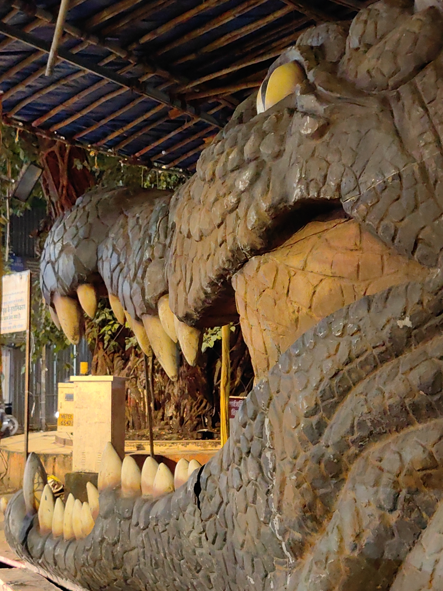 The image shows a close-up of a large, weathered sculpture of a crocodile's head, likely a prop or part of an amusement park attraction. The sculpture is made of rough, textured material, painted in shades of brown and gray, mimicking scales. Several large, pointed, yellowish teeth are visible, protruding from both the upper and lower jaws, which are open. In the background, a thatched-like roof structure made of wood and dark fabric is visible, supported by wooden beams. Behind the sculpture, greenery and trees can be seen, suggesting an outdoor or semi-outdoor setting. To the left of the sculpture, a light-colored electrical box with some text and numbers is present, along with a yellow glass. The overall impression is of a somewhat aged, perhaps somewhat kitschy, attraction. This imagery is consistent with many decorative elements found in the Kamala Nehru Park in Malabar Hill, Mumbai, India, particularly around the popular "Old Woman's Shoe" attraction, which features similar decorative elements and provides a panoramic city view.