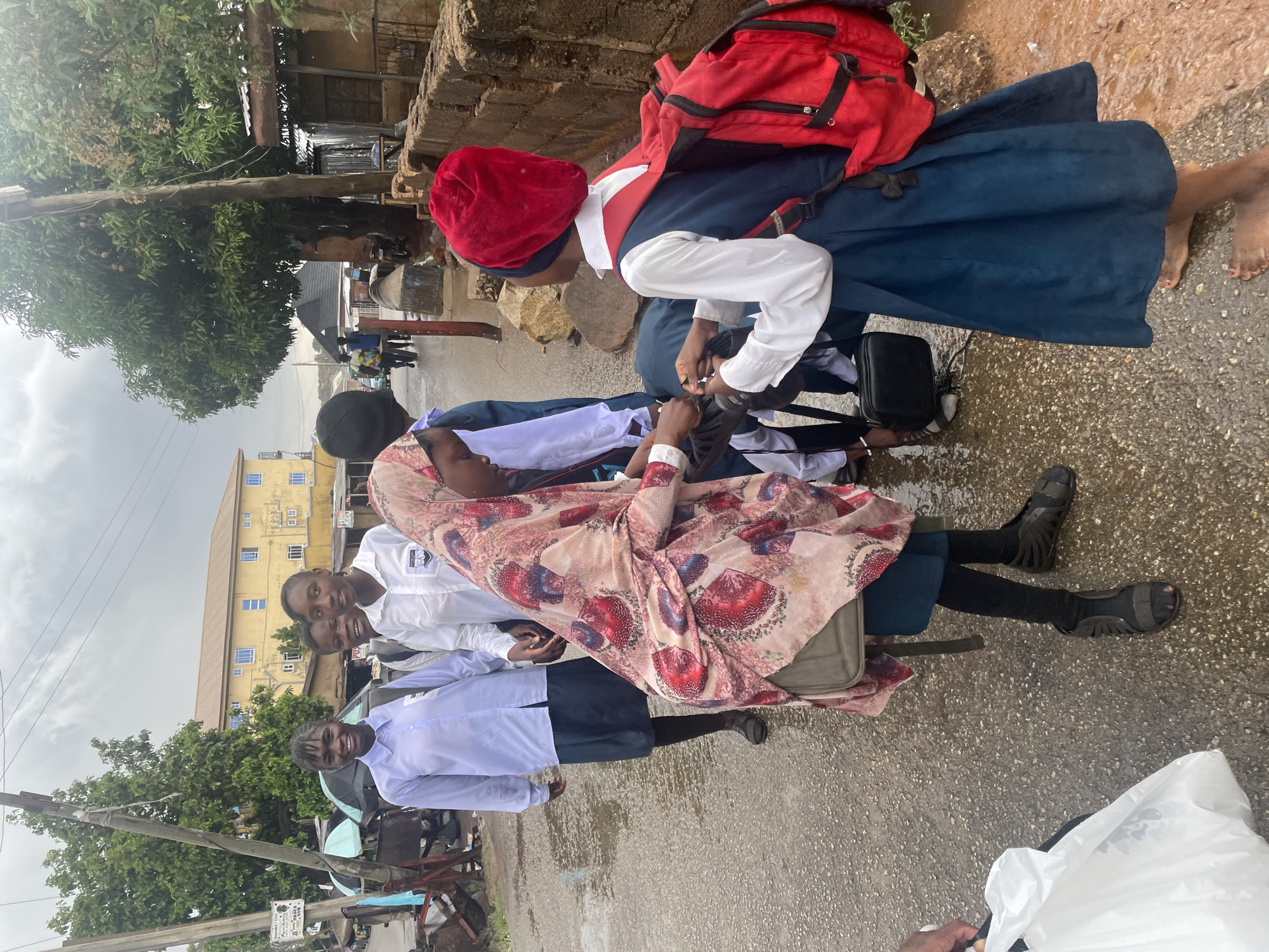 A group of African school girls stand along a wet concrete thoroughfare in Jos, Nigeria. The girls wear navy blue skirts and white, collared shirts, consistent with a school uniform. One girl wears a red head covering. A red backpack is slung over her shoulder. Another girl stands barefoot. The girls stand close together, appearing to work on an unidentified object on the ground. Other girls stand nearby, one smiling towards the camera. Utility poles, a tree, buildings, and vehicles are visible in the background, indicating an urban environment.