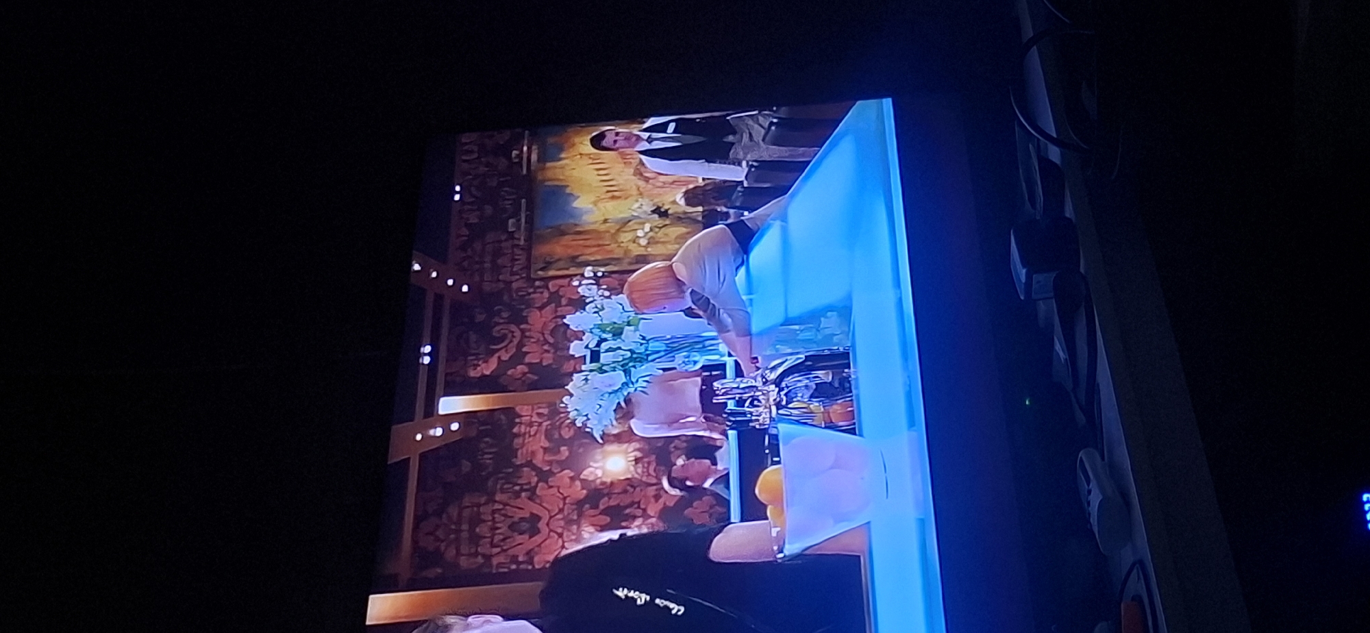 The image shows a scene from what appears to be a bar or restaurant setting. There are several people present. On the left, a person in a black shirt with white lettering on the sleeve is partially visible. In the center, a person with blonde hair is leaning over a blue counter, and behind them, a floral arrangement of white flowers is displayed in a vase. To the right of the flowers, a man in a dark suit and tie, possibly a waiter, stands and looks towards the viewer. Further back, other patrons are seated or standing, blurred by the focus. In the foreground, a glass bowl filled with oranges sits on the blue counter. The background features ornate wallpaper and artwork. There is no discernible information in the image that would indicate a location in Yerocham, Israel. The overall lighting is dim, suggesting an indoor evening setting.