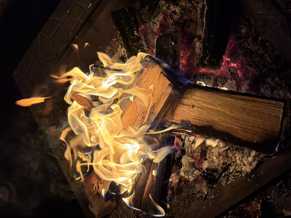 A high-angle photograph depicts an active wood fire contained within a dark, possibly metallic, fire pit or grate structure. The scene is located in Huixquilucan de Degollado, Mexico, under dark conditions, likely nighttime. Multiple wooden logs are present; some are actively burning, producing dynamic yellow-orange flames that show streaks of blue at their bases. White-grey ash and glowing red embers are visible beneath and surrounding the burning wood, indicating significant combustion. Faint trails of smoke are observed rising from the flames against the dark background. No individuals, objects beyond the fire pit and its contents, or identifiable interactions are visible. The fire itself is the primary light source, illuminating the immediate area.