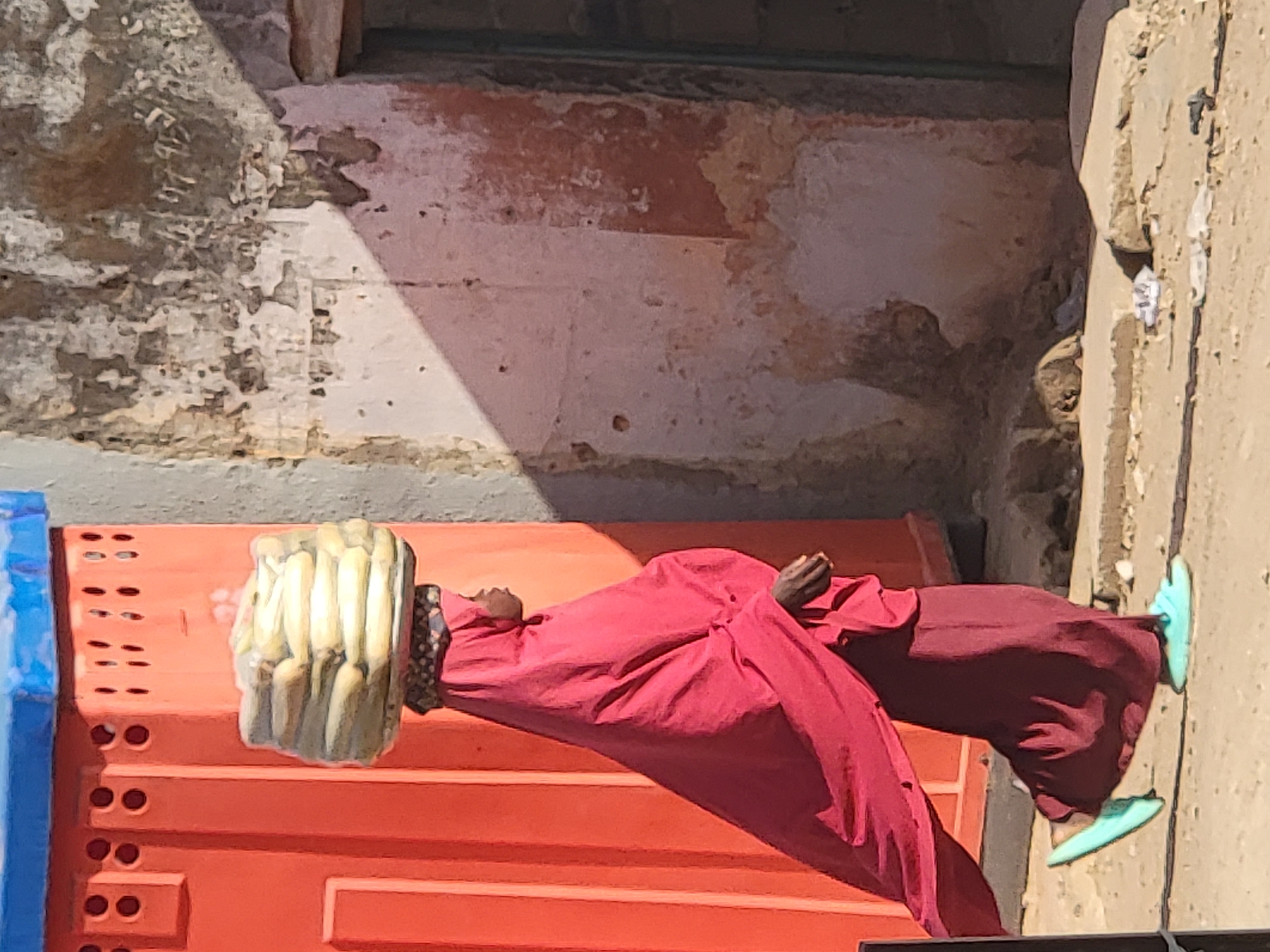 A person lies on an orange structure on a sidewalk in Maiburiji, Nigeria. The person is wearing a red garment and light blue shoes, with a large, layered headpiece that appears to be crafted from dried material. The person's hands are clasped in front of them. The orange structure is next to a blue object and is positioned against a weathered wall. The sidewalk and wall are textured with cracks and discoloration.