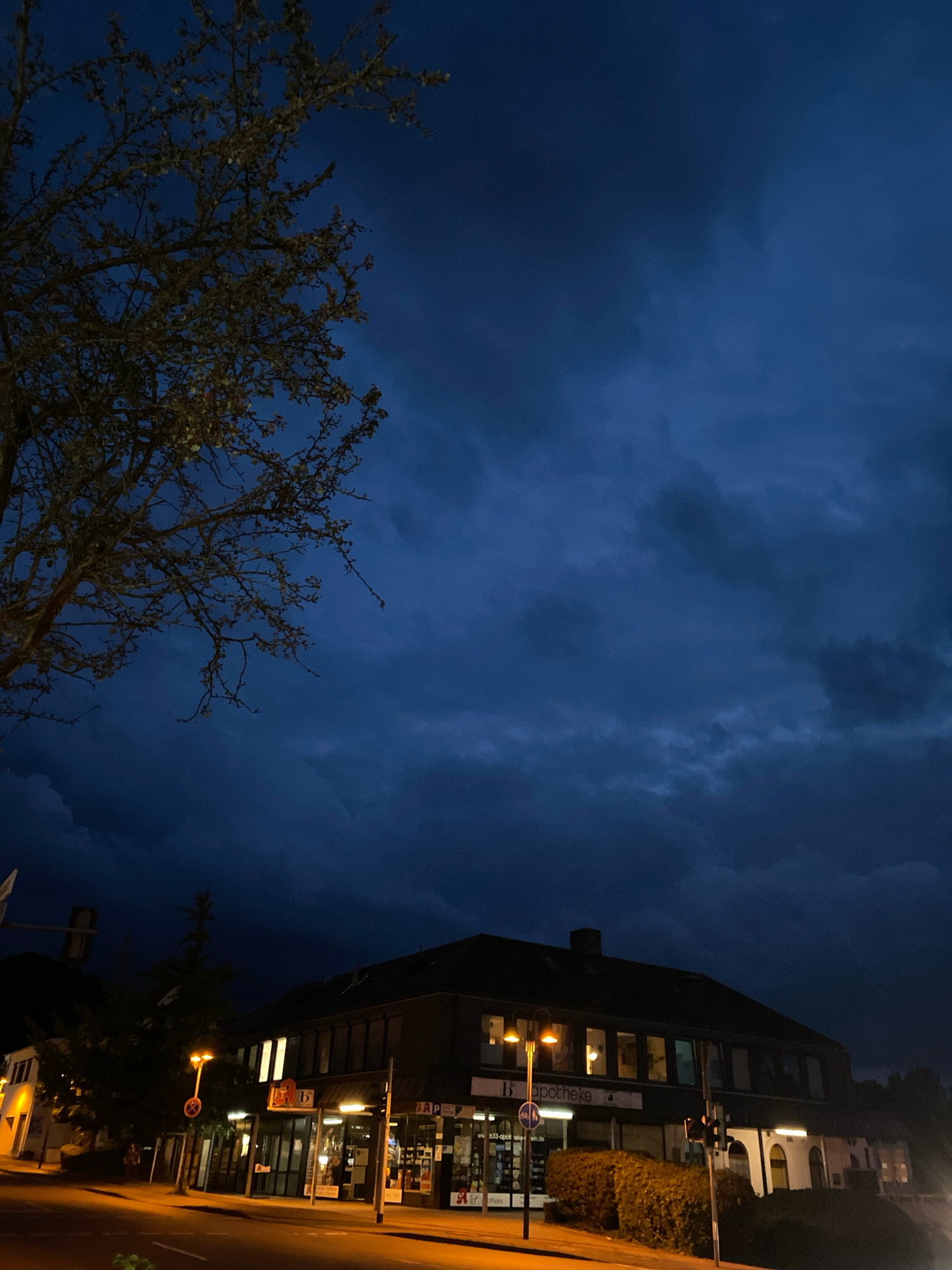 The image depicts a nighttime scene in Rinteln, Germany. In the foreground, a street intersection is visible with streetlights illuminating the road and sidewalks. To the right, a dark-colored building houses businesses with lit windows and signage. One sign clearly reads "Apotheke," indicating a pharmacy. A bare-branched tree is silhouetted against a dark, cloudy sky in the upper left quadrant of the image. The overall atmosphere is dim and somewhat ominous due to the dark sky and the limited light sources. No individuals are clearly discernible in the scene.