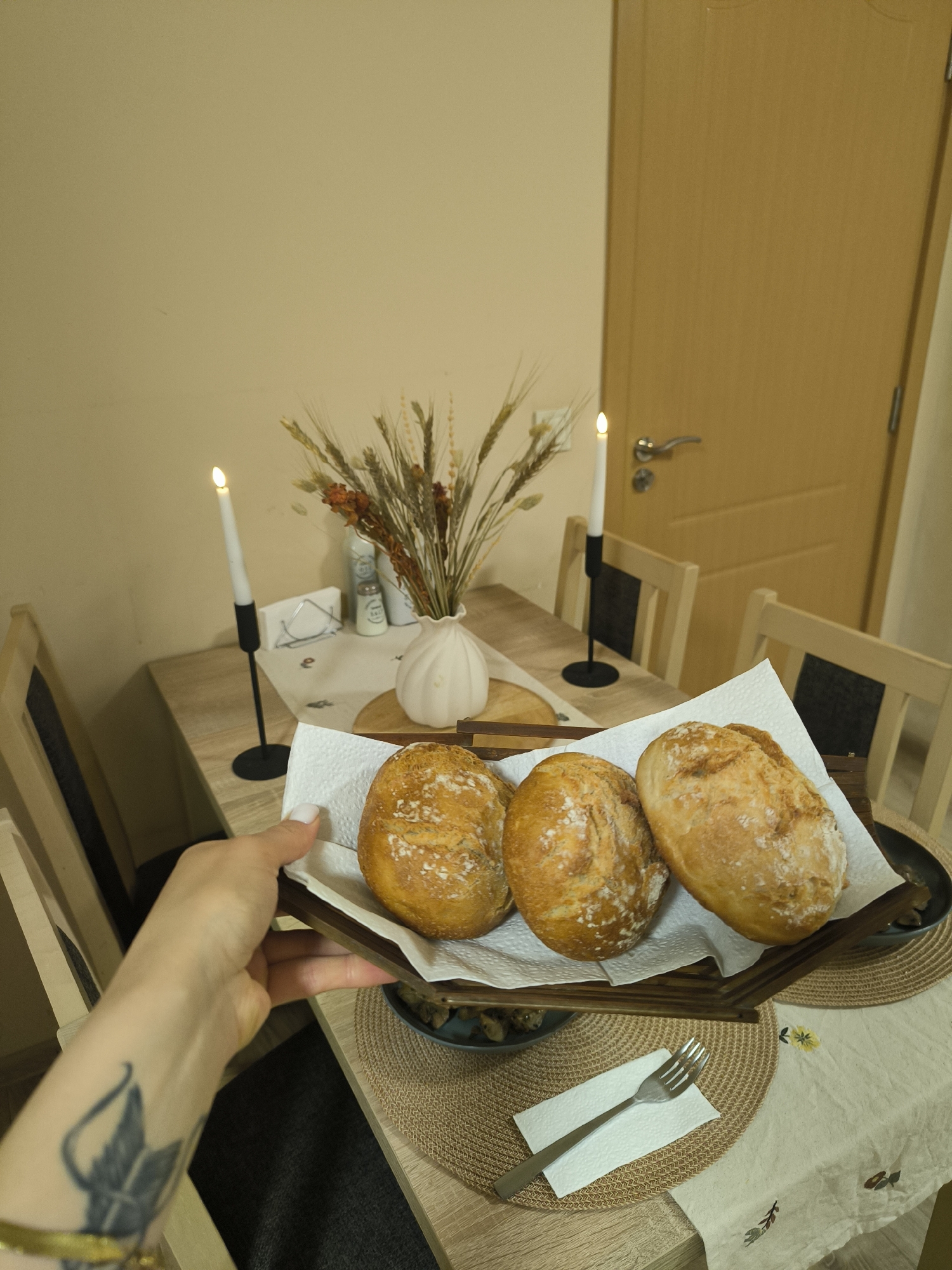An indoor scene depicts a person's left arm and hand holding a dark wooden tray with three golden-brown bread rolls, each dusted with a white powder, resting on white paper towels. The hand has white nail polish on the index finger and a black tattoo resembling a stylized leaf or feather on the forearm, along with a golden bracelet on the wrist. The tray is positioned over a light brown woven placemat on a light wooden dining table. To the right, another similar placemat holds a silver fork on a white paper napkin. Adjacent to this placemat, a cream-colored tablecloth with embroidered yellow and brownish floral motifs is visible.
The table is set with two lit white taper candles in slender black metal holders, creating an ambient glow. A white, textured, garlic-shaped ceramic vase containing a dried arrangement of wheat stalks and reddish-brown flora serves as a centerpiece. Behind the vase, a small wooden trivet or cutting board is visible. Other items on the table include a white rectangular napkin holder, two small cylindrical shakers, and a blue spray bottle. Two light wooden dining chairs with dark fabric inserts in their backs are partially visible on either side of the table. The background features a plain light beige wall and a light brown wooden door with a silver handle, slightly ajar on the right. The overall location is an Unknown location.