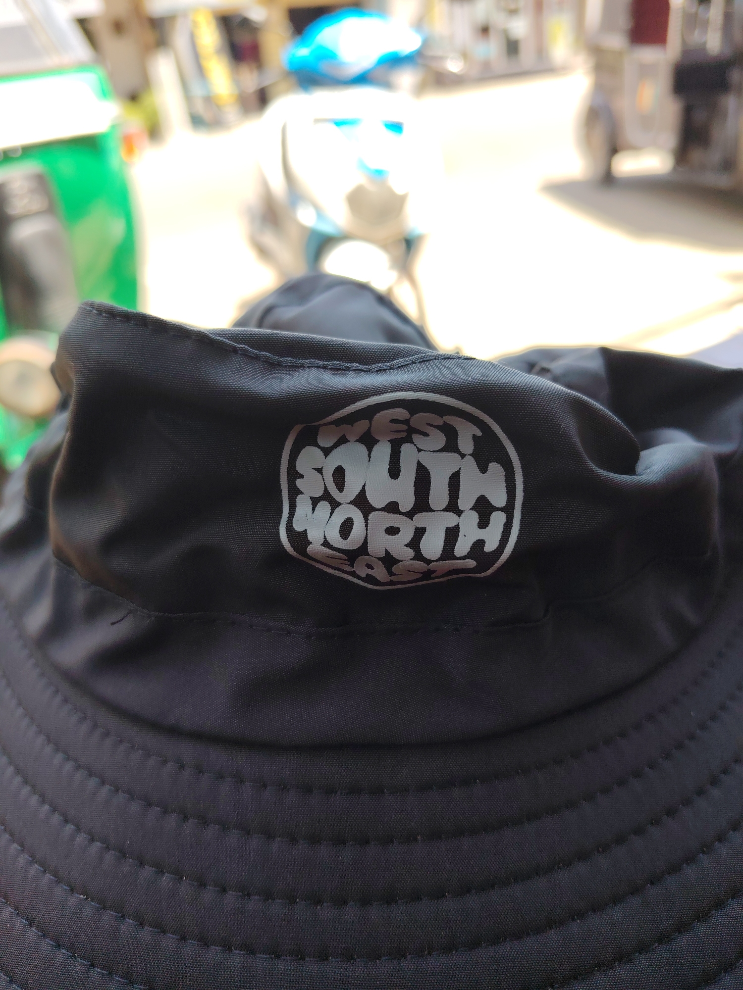 A close-up, eye-level shot shows a black bucket hat in the foreground. The hat has a circular white logo on it with text that appears to read "WEST SOUTH NORTH EAST." The background is out of focus, showing a street scene with a green vehicle on the left, and various structures and possibly other vehicles in the distance. The lighting suggests it is daytime.