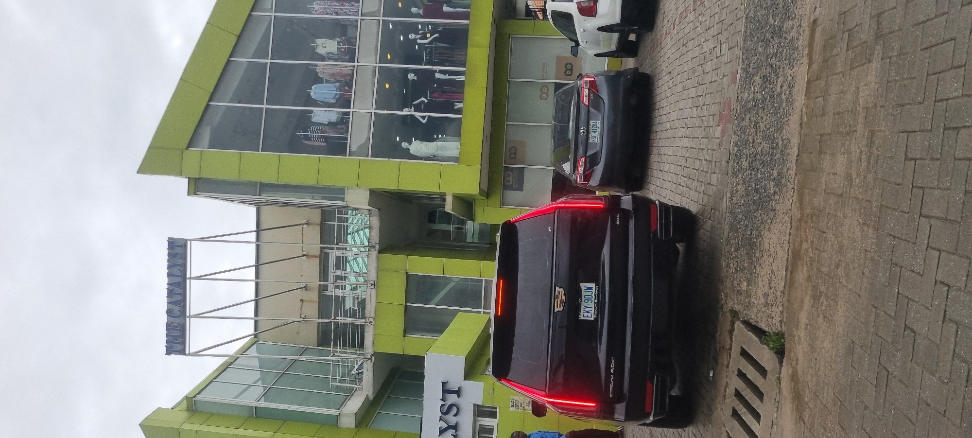 The image shows the front of a modern, lime-green building with large glass windows. A sign above the entrance reads "THE CATALYST." In front of the building, there are three cars parked on a cobblestone pavement. The car in the foreground is a black Cadillac Escalade with its brake lights illuminated. To the right of the Escalade is a dark grey Toyota Camry, and to the right of the Camry is a white SUV. A person in a blue shirt stands to the left of the Escalade. The building appears to be a commercial establishment, possibly a retail store, with mannequins visible inside the windows. The sky is overcast. The scene is located in Moba, Nigeria.