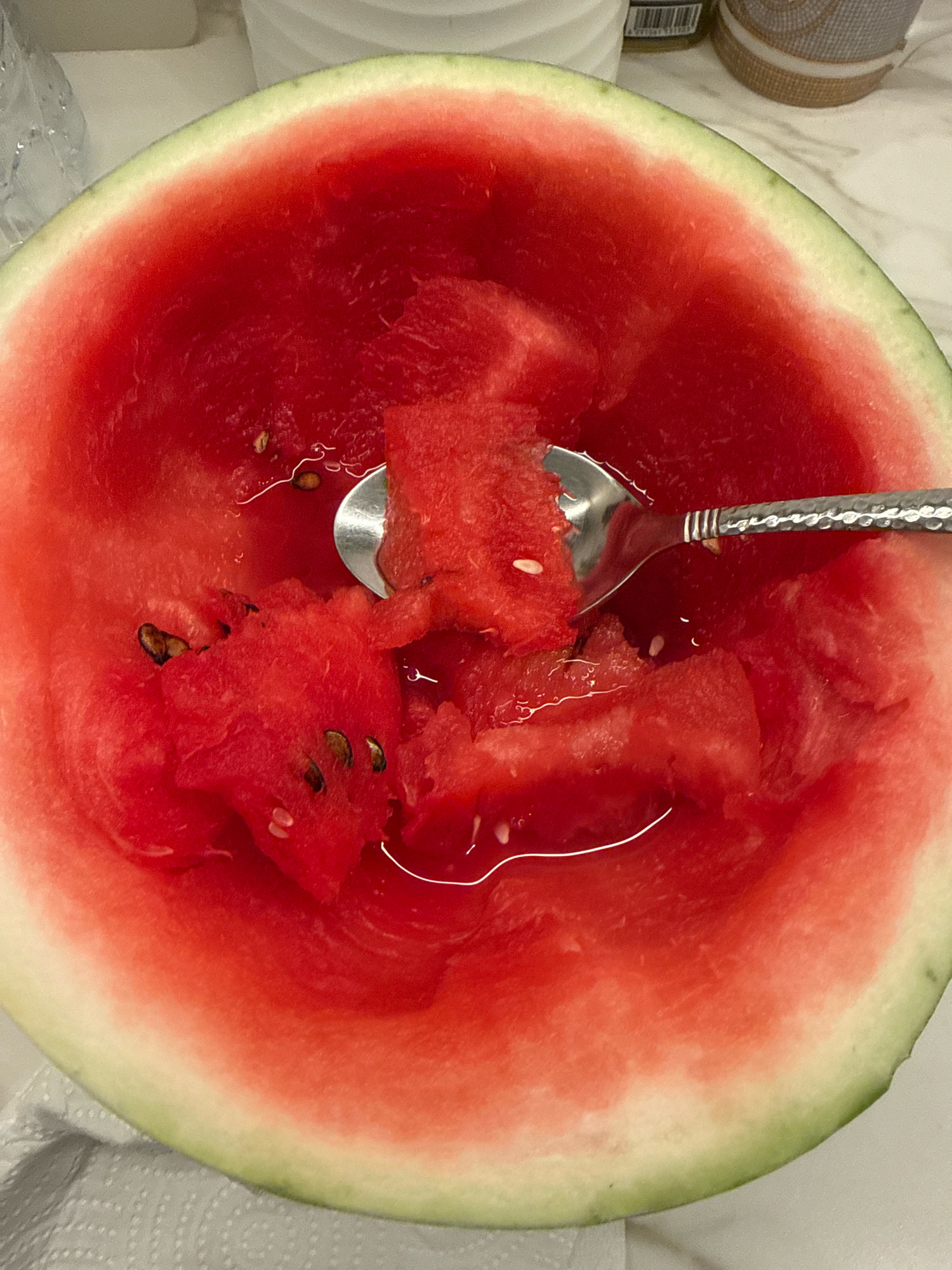 A close-up, overhead shot shows a half of a watermelon that has been hollowed out and is filled with chunks of watermelon flesh. A metal spoon with a textured handle is lodged in the watermelon, scooping out a piece of flesh. There are several black watermelon seeds scattered throughout the flesh and juice. The rind of the watermelon is visible around the edges, showing a transition from green to white to pink. The watermelon is sitting on a light-colored, speckled surface, possibly a marble countertop, with a white paper napkin visible in the bottom left corner. The background is blurred but appears to include some household items, including a partially visible bottle and a decorative object. There is no indication of the location being Dubai, United Arab Emirates.