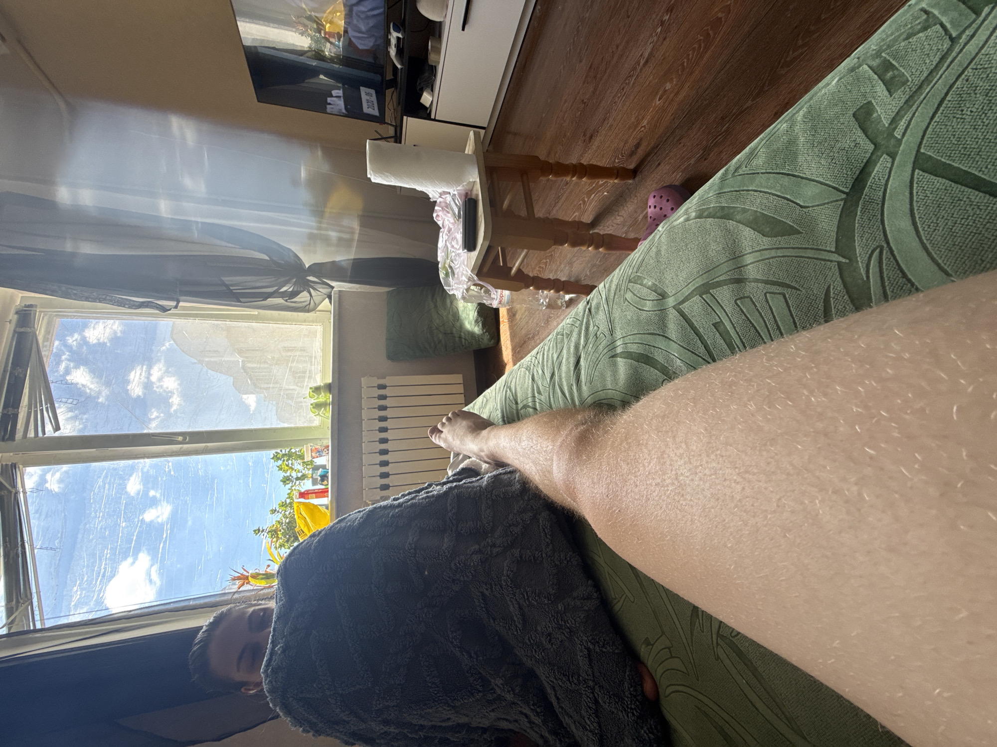 The image shows a first-person view of a person's leg resting on a green couch. To the left, a dark gray blanket covers another person's torso and head, with only their eyes and forehead visible. In the background, a large window looks out onto a bright sky with clouds and buildings. A radiator is visible below the window. To the right, a small wooden stool holds a roll of paper towels, a phone, and some fabric. A television is mounted on a white cabinet further to the right. The floor is made of polished wood. A pink slipper is visible on the floor near the couch. The scene appears to be an indoor living space.
