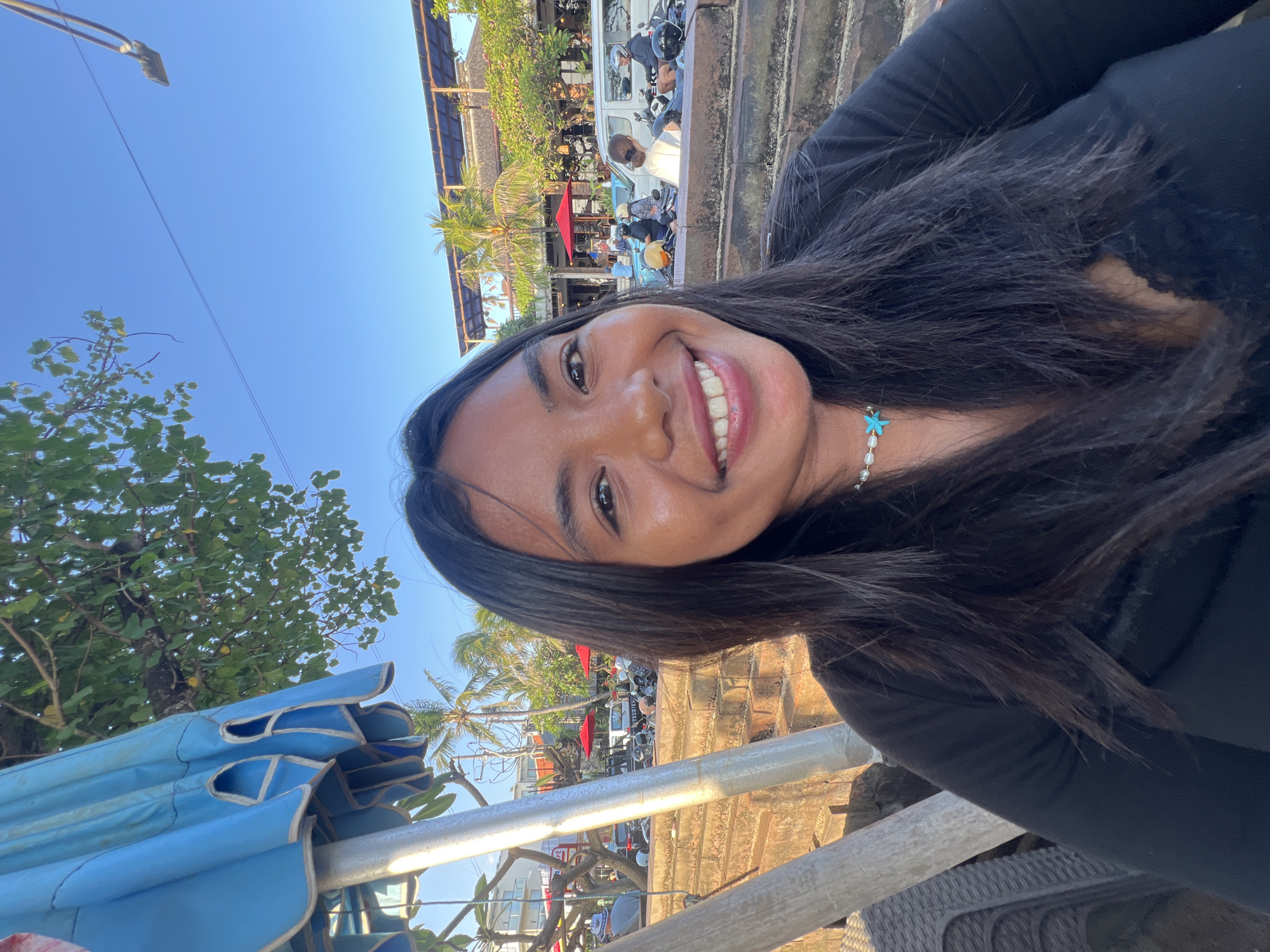 A young woman with long, dark hair and a bright smile is taking a selfie outdoors. She is wearing a black long-sleeved shirt and a necklace with pearls and a blue star charm. Behind her, a large blue umbrella is partially visible, along with palm trees, buildings, and a clear blue sky. The background suggests a bustling outdoor area, possibly a beachfront cafe or market in Kuta, Indonesia, given the presence of scooters and tropical vegetation. The lighting indicates it is daytime, likely late afternoon due to the warm tones.