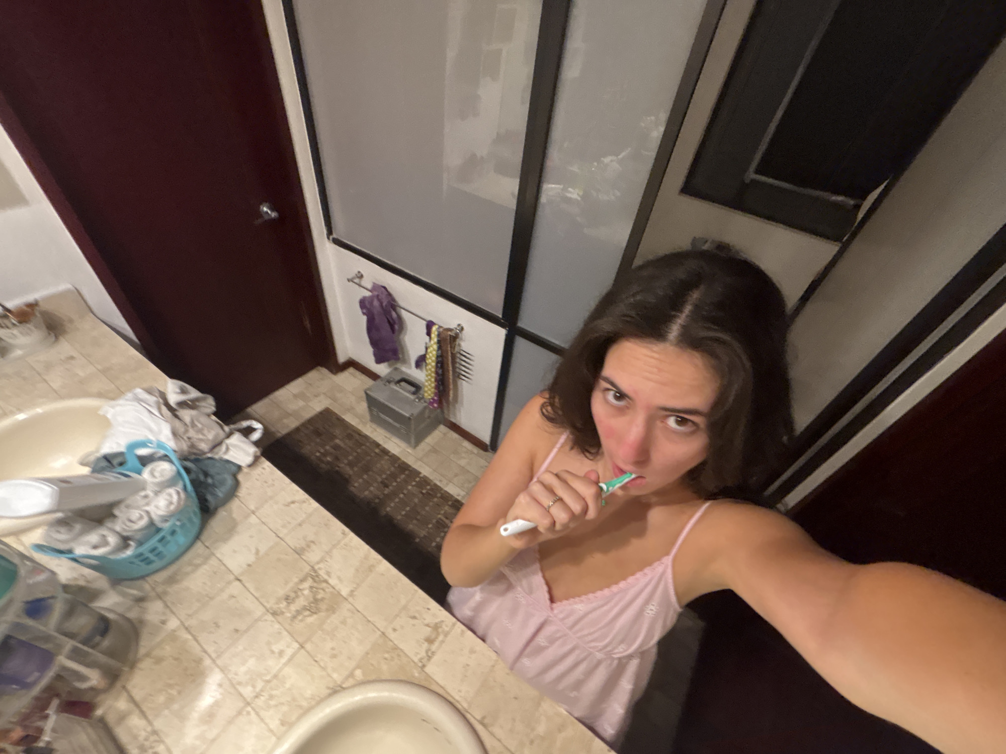 A young woman is taking a selfie in a bathroom. She is holding a toothbrush in her mouth and has dark hair. She is wearing a pink, floral-patterned camisole. The bathroom has light-colored tile countertops and floors. There is a sink and a laundry basket filled with towels on the counter. A rug is on the floor near a gray storage box. A towel rack with towels hangs on the wall. A door is visible on the left. The location is not apparent from the image.