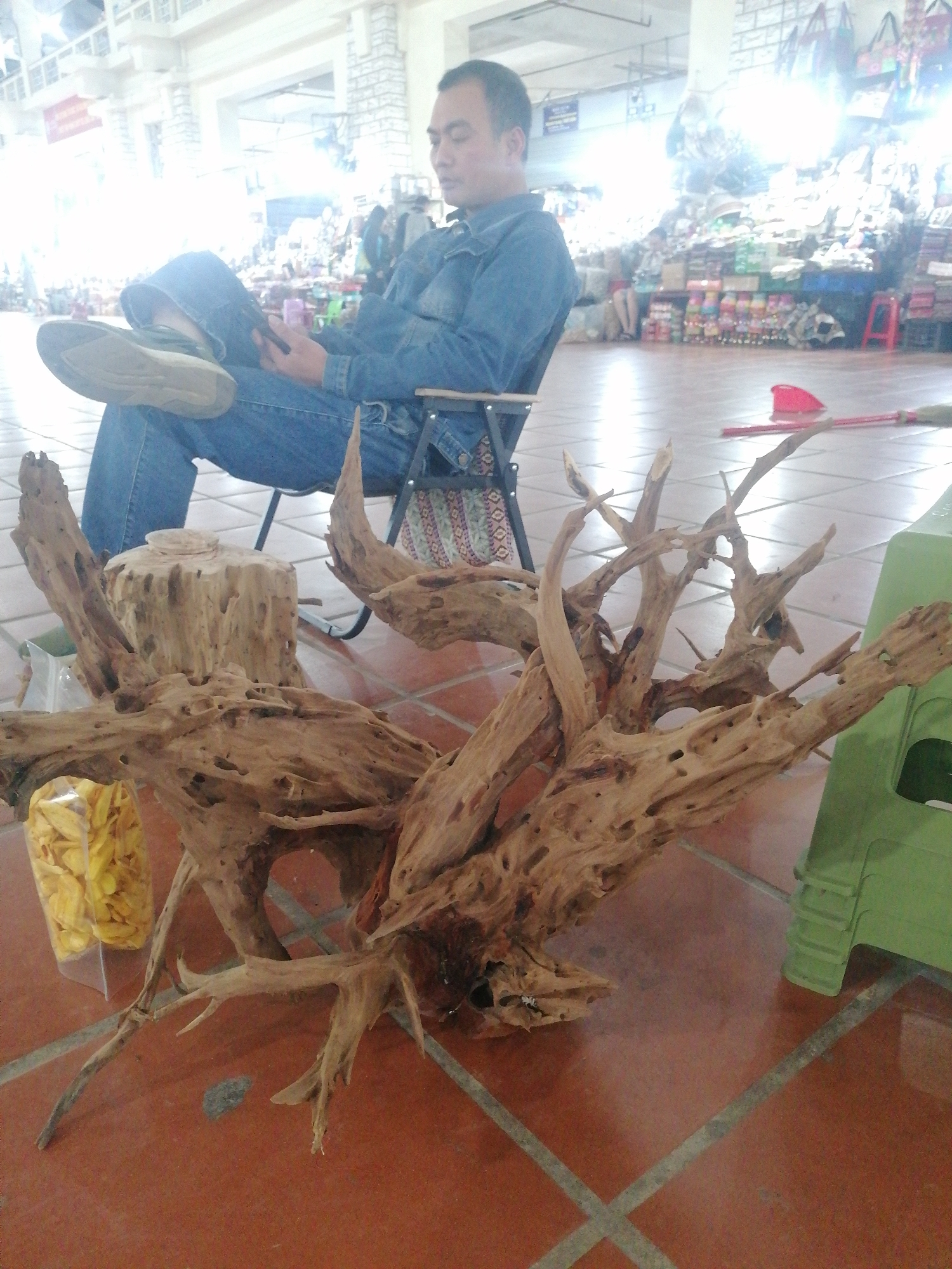 This image depicts a scene inside an indoor market in Sa Pa Ward, Vietnam.
In the foreground, a large, gnarled piece of light brown driftwood or root system rests on a tiled floor. The floor is covered with reddish-brown tiles separated by grey grout lines. To the left of the driftwood, a clear plastic bag containing yellow, dried fruit slices (possibly jackfruit or banana chips) is visible. Behind the main driftwood, a smaller, cylindrical wooden stump stands. On the right side of the driftwood, a green plastic stool or container is placed on the floor. Further back on the floor, a red plastic dustpan and part of a broom handle are present.
A man is seated in the midground, occupying a black metal folding chair. He wears a blue denim shirt and blue jeans, with his left leg casually crossed over his right. He has short, dark hair and appears to be looking down at an object in his hands, possibly a mobile phone. He seems relaxed, suggesting he is resting or waiting.
The background reveals numerous market stalls displaying various goods, though they are somewhat overexposed and blurry, making specific items indistinct. The bright lighting in the background suggests it is daytime. The overall setting is typical of a bustling market, even if the immediate foreground activity is calm. There is a red sign with unreadable text in the far background due to the bright lighting.