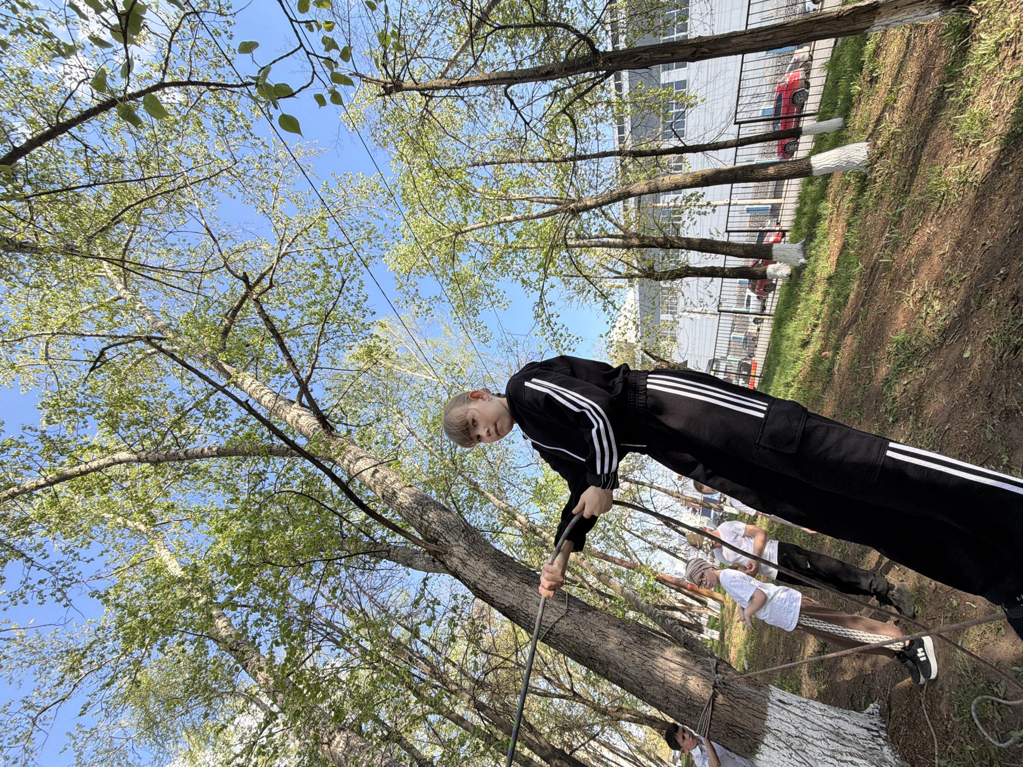 A young person in a black tracksuit with white stripes is participating in a rope course in Izhevsk, Russia. The course is set up between trees with white-painted trunks. Other individuals are present, some wearing white shirts. A building with a metal fence and a red car parked nearby are visible in the background. The sky is blue with scattered clouds.