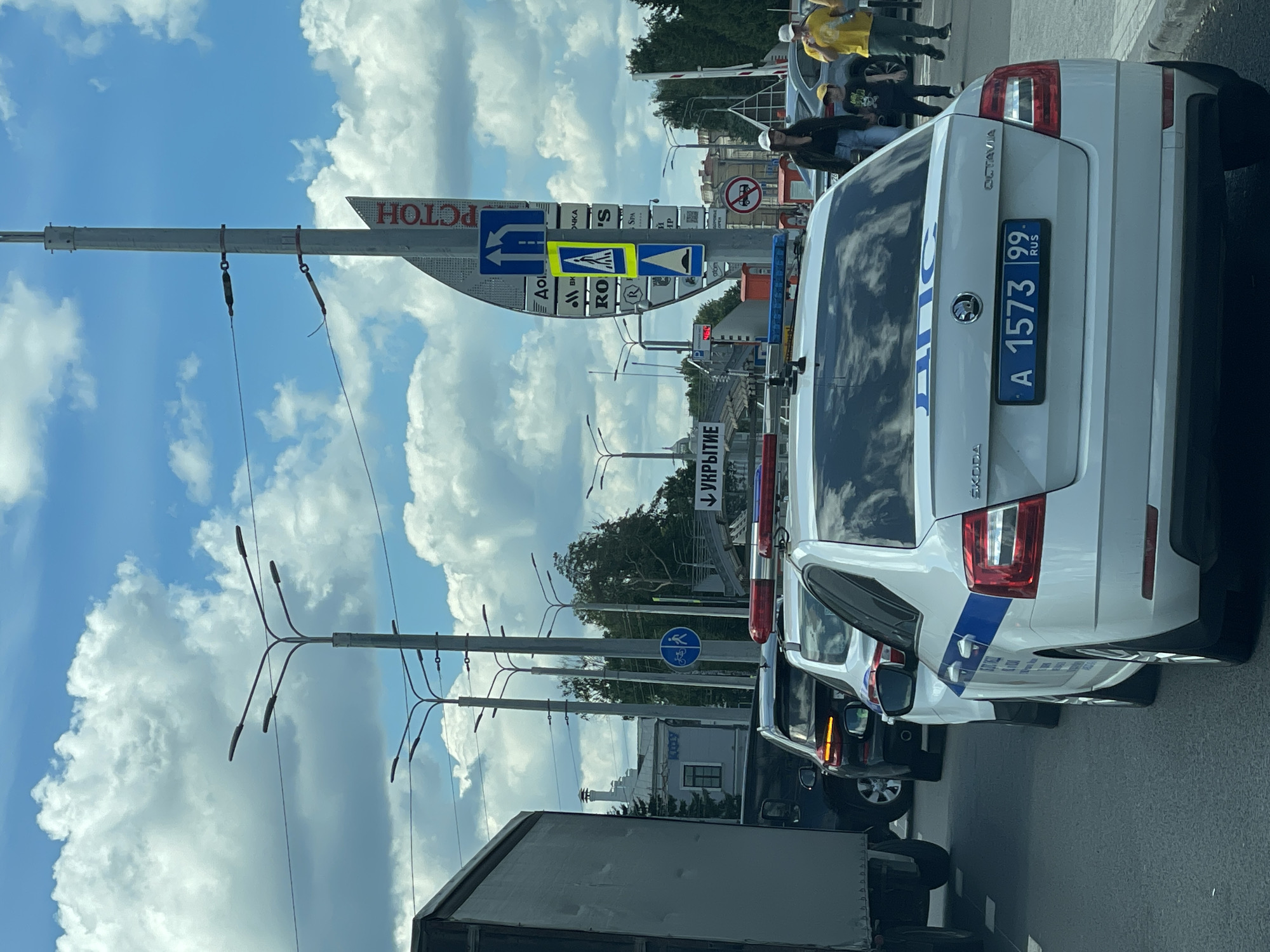 The image depicts a traffic scene in Kazan, Russia, on a sunny day with scattered clouds. The foreground is dominated by the rear of a white police car, identified by its blue and white markings and the Cyrillic text "ДПС" on its trunk. The license plate reads "A 1573 99 RUS". To the left of the police car, part of a large grey truck is visible. Behind the police car, other vehicles are stopped in traffic, including a dark SUV with its brake lights illuminated and another white car.
In the background, several tall lampposts with multiple lights are prominent. A traffic signpost displays a combination of directional signs and a pedestrian crossing symbol. On the right side of the frame, two people are walking on the sidewalk. One is a woman in a white cap and dark clothing, and the other is a boy in a yellow t-shirt and a dark cap. Further back, a man in a yellow t-shirt and dark pants is also visible. Beyond the traffic and signs, buildings and trees can be seen. The overall impression is of a typical urban traffic situation.