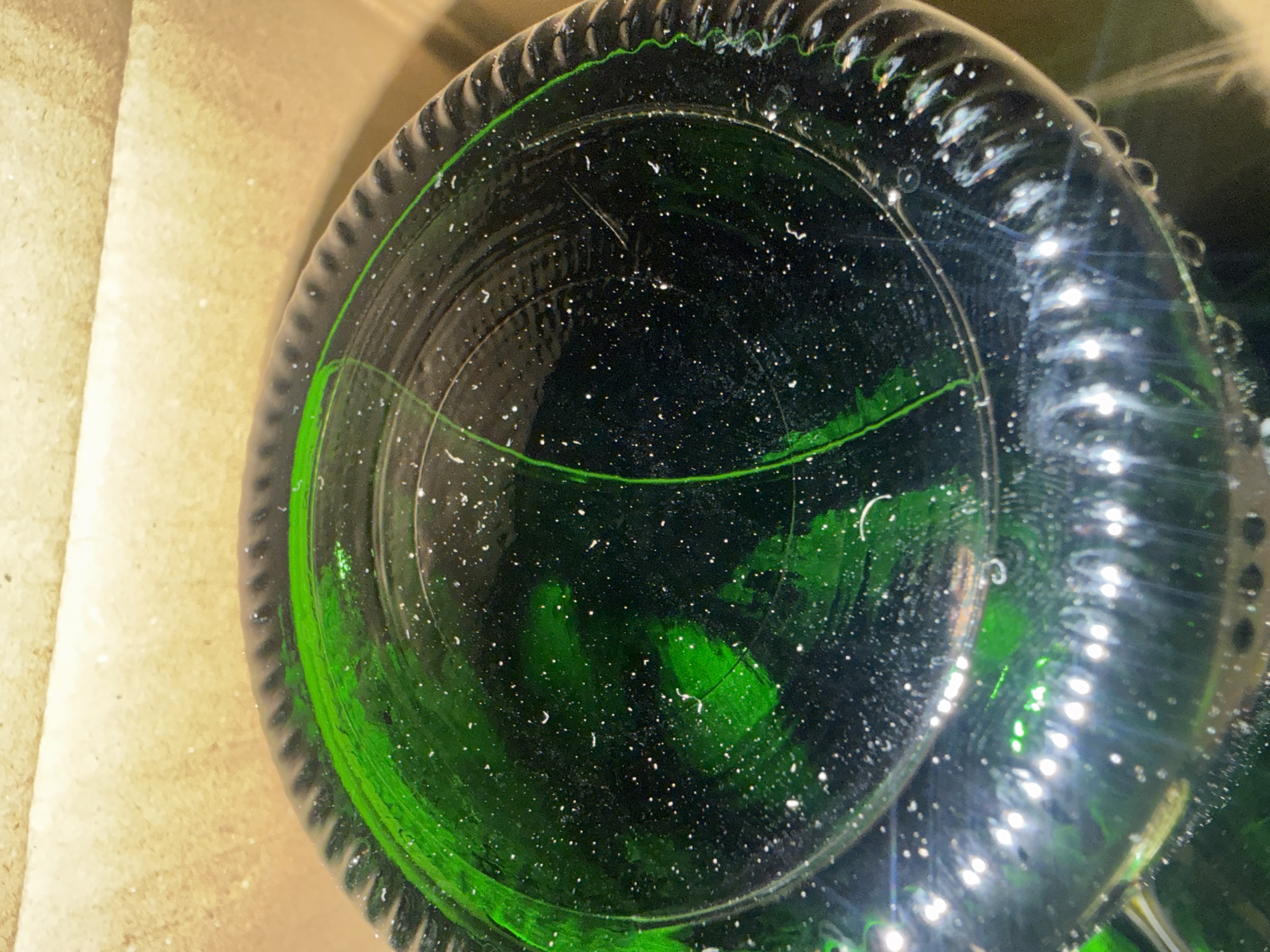 An extreme close-up shot, looking down into the bottom of a green glass bottle. The bottom of the bottle has a ribbed texture around the edge and a slightly concave surface. The glass is speckled with many tiny white dots, appearing as if it has been frosted or is dirty. Streaks of brighter green color are visible within the bottle, suggesting a liquid residue or coating. The bottle is placed on a textured, light brown surface, possibly cardboard. Reflections of bright light sources are visible on the curved surface of the bottle at the bottom of the image, creating a shimmering effect. The overall impression is one of a close examination of the interior of a glass container.