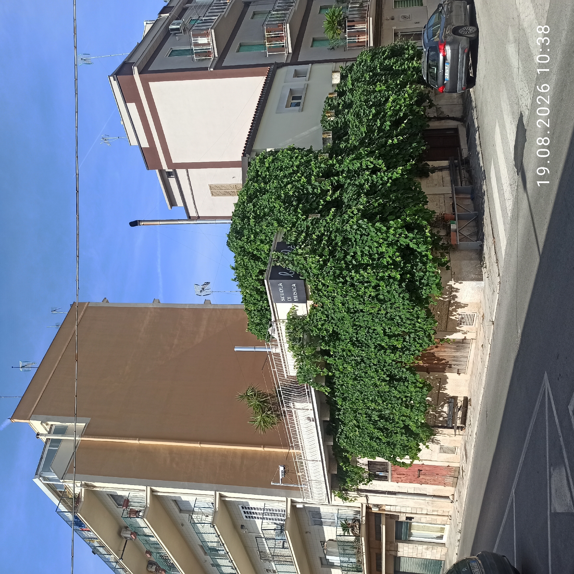The image shows a street scene in Ruvo di Puglia, Italy, on August 19, 2026, at 10:38 AM. The scene features a corner intersection with multiple buildings and a dark gray station wagon parked on the right side of the road.
In the foreground, a street with a pedestrian crossing is visible, with shadows cast by the sun. A dark gray station wagon, license plate FD 575LB, is parked at the right edge of the frame, facing away from the viewer.
The buildings on the left and in the background are multi-story structures. The building on the left is a light-colored apartment building with multiple balconies. The building in the center is a taller, light brown building. To the right of the light brown building, a smaller building with a cream-colored facade and brown trim is visible. A large amount of lush green ivy covers a portion of the lower levels of the buildings, particularly around the center and right side. A sign on the ivy-covered section reads "SCUOLA DI MUSICA." There is a white railing with decorative elements on a balcony area above the ivy.
The environment is sunny with a clear blue sky. Power lines are visible strung across the sky. There are several antennas on the rooftops of the buildings. A metal bench is situated on the sidewalk area to the left of the parked car, partially obscured by the ivy.