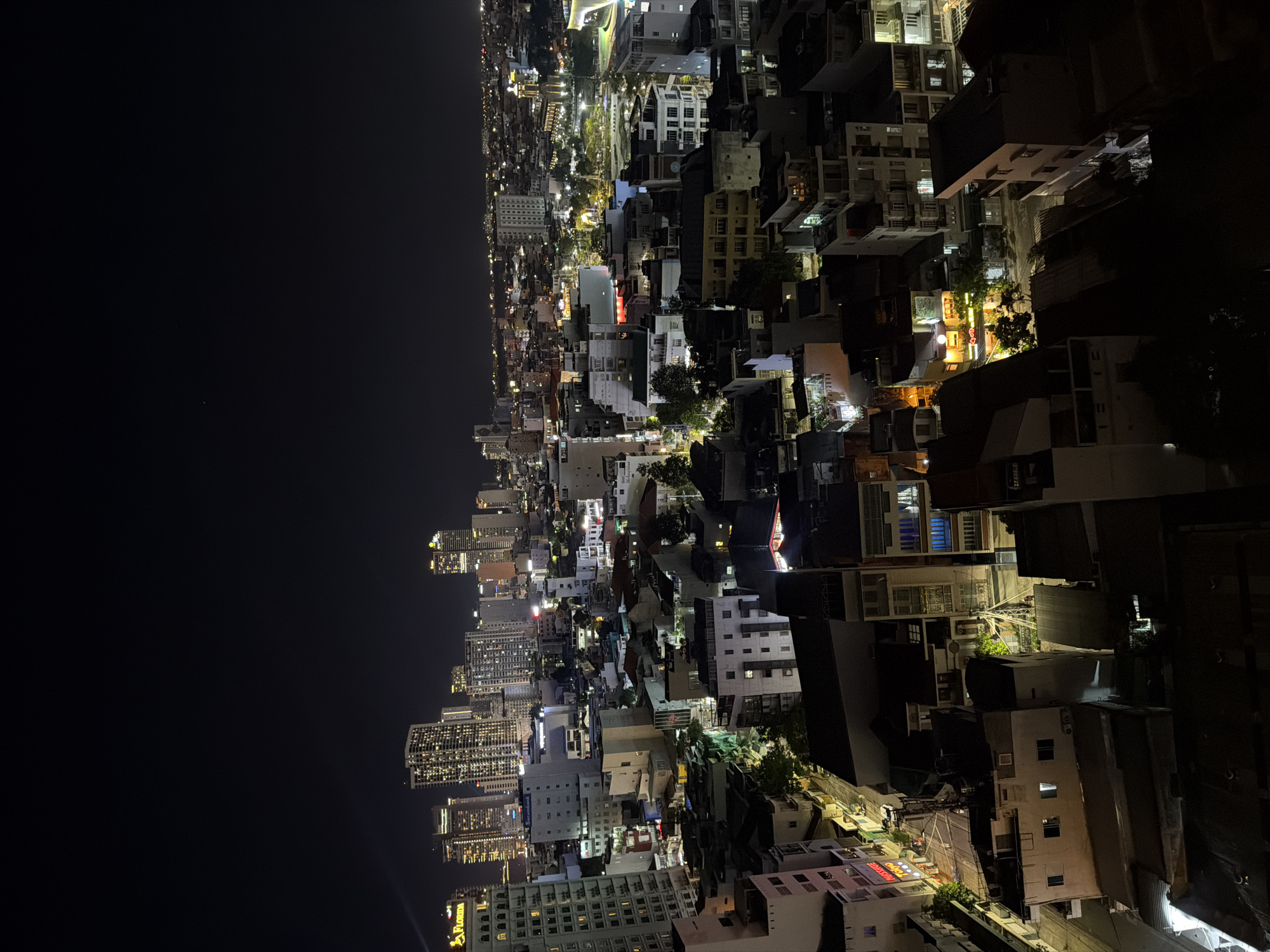 This is an aerial nighttime photograph of a densely built urban environment, likely located in Nha Trang Ward, Vietnam. The image captures a sprawling cityscape illuminated by artificial lights, with numerous buildings of varying heights and architectural styles. Tall, modern high-rise buildings with illuminated windows are visible in the background, contrasted by lower, older structures in the foreground. The scene depicts a network of streets and alleyways winding between buildings, with some areas appearing residential and others commercial, indicated by illuminated signs and storefronts. Vegetation, such as trees, can be seen interspersed among the buildings, some with lights emanating from within. The sky is dark and clear, with the absence of visible stars or celestial bodies. There are no people or vehicles clearly discernible in the image, suggesting a general overview of the urban landscape at night. The lighting creates a contrast between illuminated interiors and darkened exteriors, highlighting the textures and forms of the urban architecture.