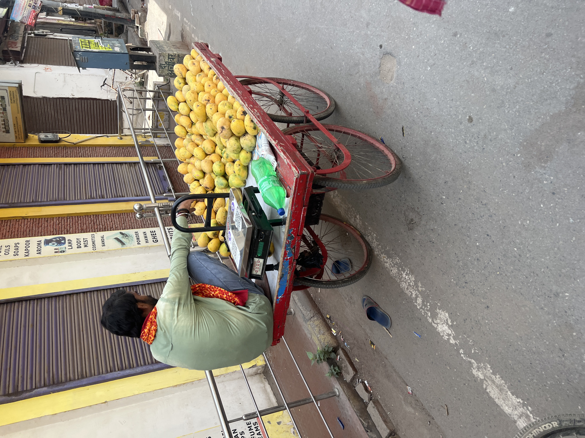 A street vendor sits on a tricycle cart loaded with mangoes, likely selling them to passersby in Varanasi, India. He wears a green shirt and a red scarf around his neck. The cart is red with three bicycle wheels, and a green plastic bottle rests among the fruit. He is positioned on the edge of the street, next to a building with a yellow and gray facade. The building has a sign advertising various products like "OILS, SOAPS, KAPOOR, AROMA, LAMP, BODY, MIST, COSMETICS, SANDAL, POWDER, HAWAN, GHEE" and another sign advertising "9140579434, 7081990903" at a window. The street surface is asphalt, with visible debris and wear. A single blue sandal lies on the ground. A railing separates the cart from the building.
