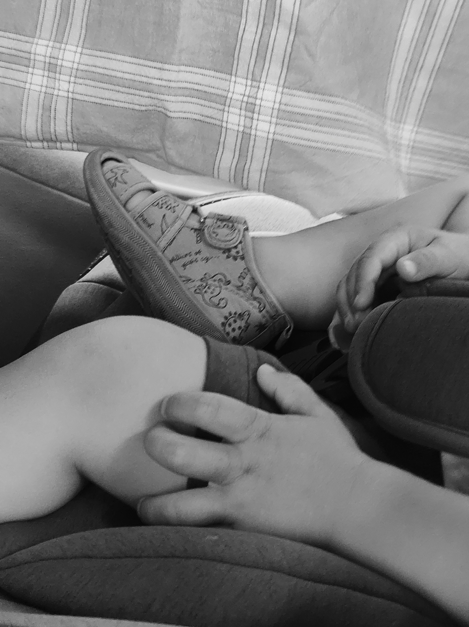 The image depicts a close-up, black and white view of a child's legs and feet. The child is wearing a patterned shoe. One foot is resting on the other leg, which appears to be the child's knee. The child's hands are also visible, one appearing to scratch the leg. The background consists of a striped fabric surface, likely a blanket or cushion. The setting is likely an indoor domestic environment. There are no discernible landmarks or features to identify a specific location in Chaikovsky, Russia.